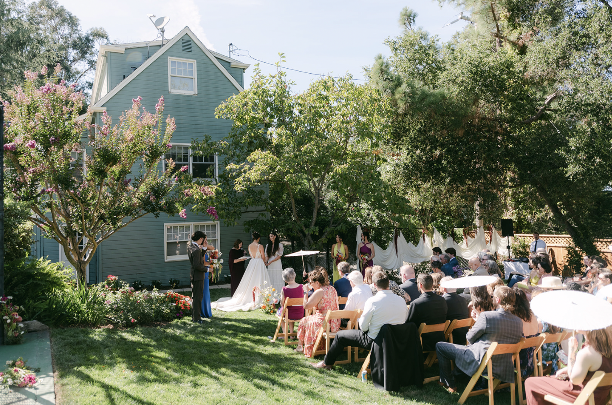 Outdoor wedding ceremony in a residential backyard with guests seated on wooden chairs, the bride and groom standing under a white fabric drape, surrounded by trees, flowers, and a blue house.