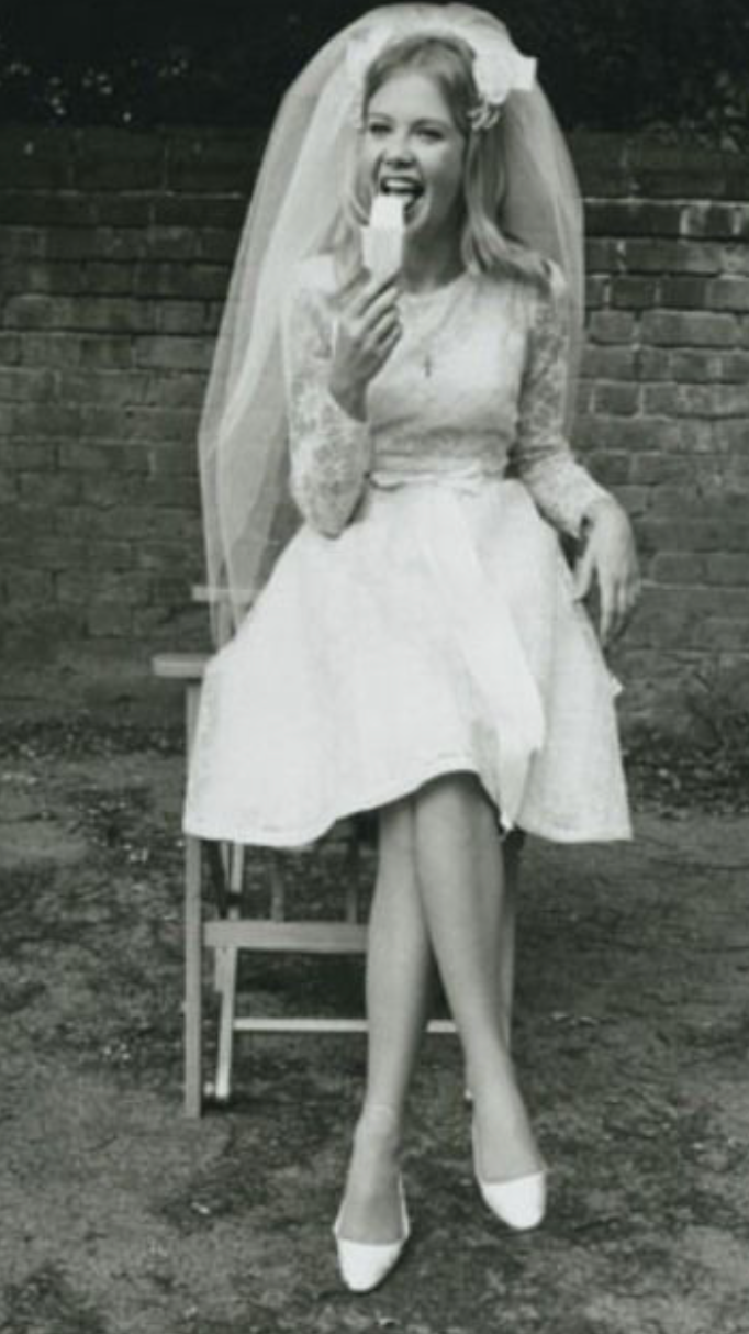 A woman in a wedding dress sitting on a chair outdoors, wearing a veil and a lace top, biting into an ice cream bar, smiling, with a brick wall in the background.