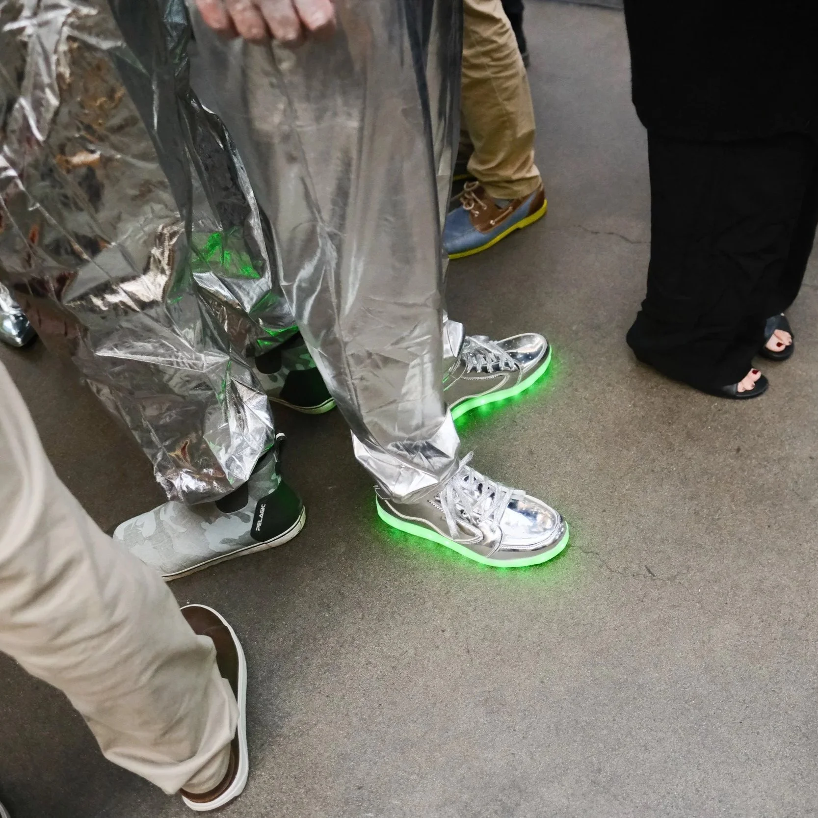 People standing indoors, wearing fashionable shoes, including shiny silver sneakers with neon green lights on the soles, and casual footwear. One person is in silver reflective clothing, and a few others are visible in the background.