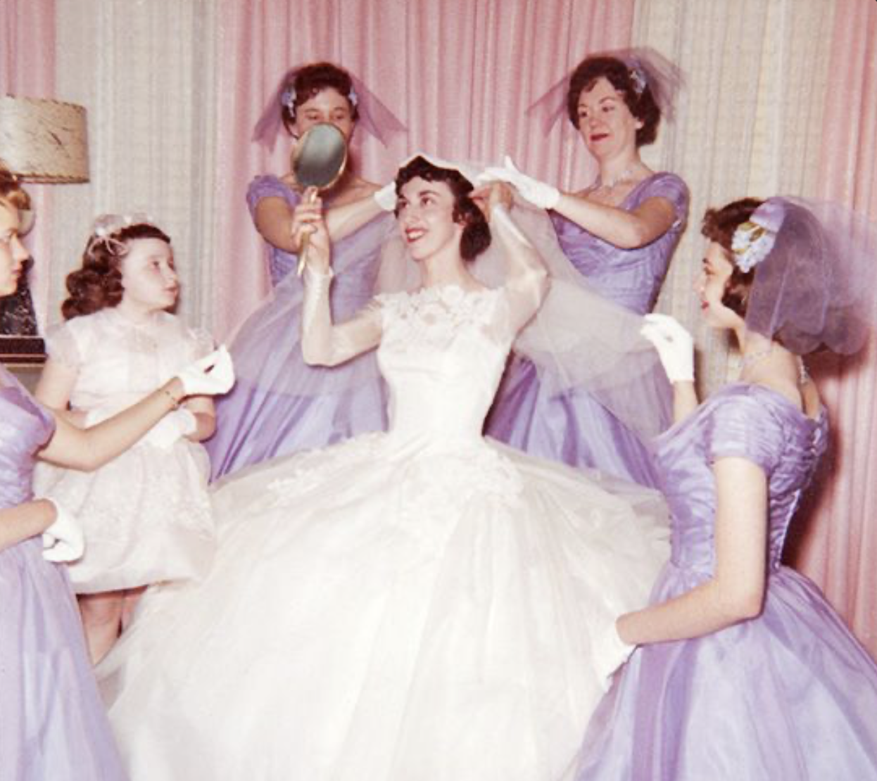 A woman in a wedding dress surrounded by five women and two young girls, all dressed in purple gowns, preparing her for a wedding or photo shoot in a vintage setting.