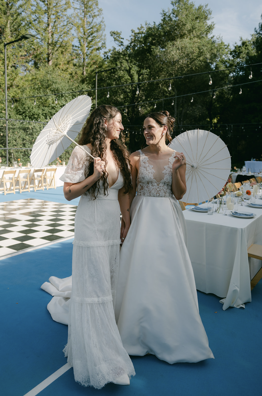 Two women in wedding dresses holding white parasols and smiling at each other, outdoors at a wedding reception with decorated tables and string lights.