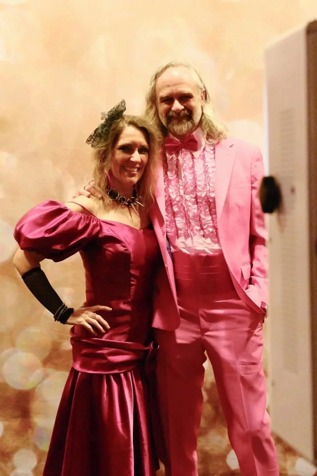 A woman and a man dressed in pink formal attire, posing together inside a room with beige walls.