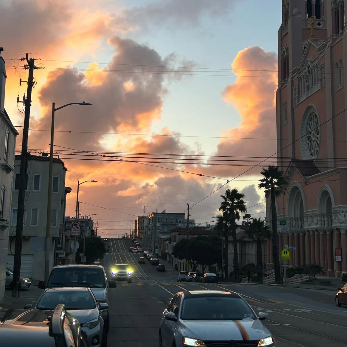 Street view during sunset with clouds, parked cars, palm trees, and a pink building with ornate architecture.