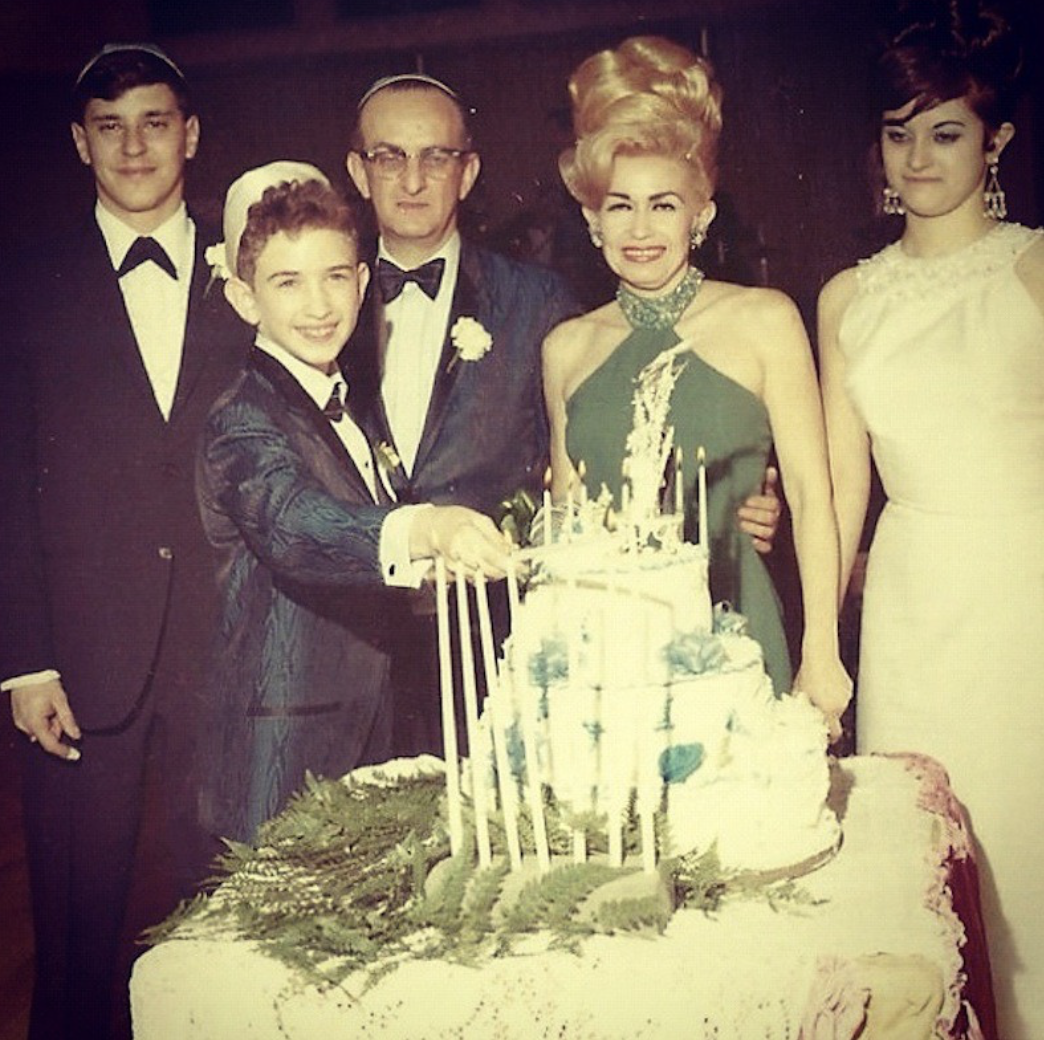 A family celebrating a special occasion with a birthday cake, five people dressed in formal attire, including a young boy, two women, and two men, all smiling.