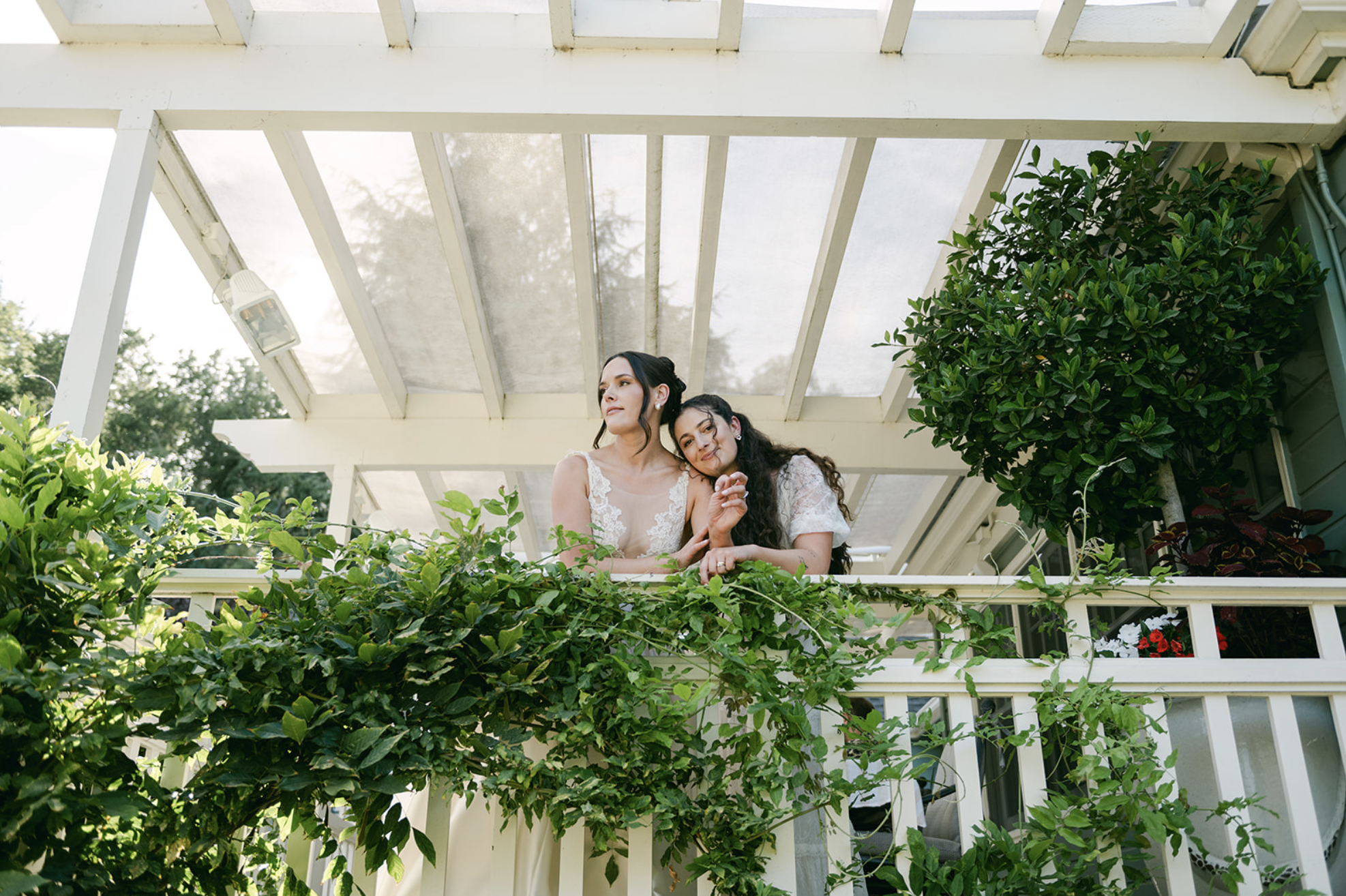 Two women, one in a white dress and the other in a white lace top, are standing on a balcony surrounded by greenery. They are looking out and smiling, with a glass roof structure above them.