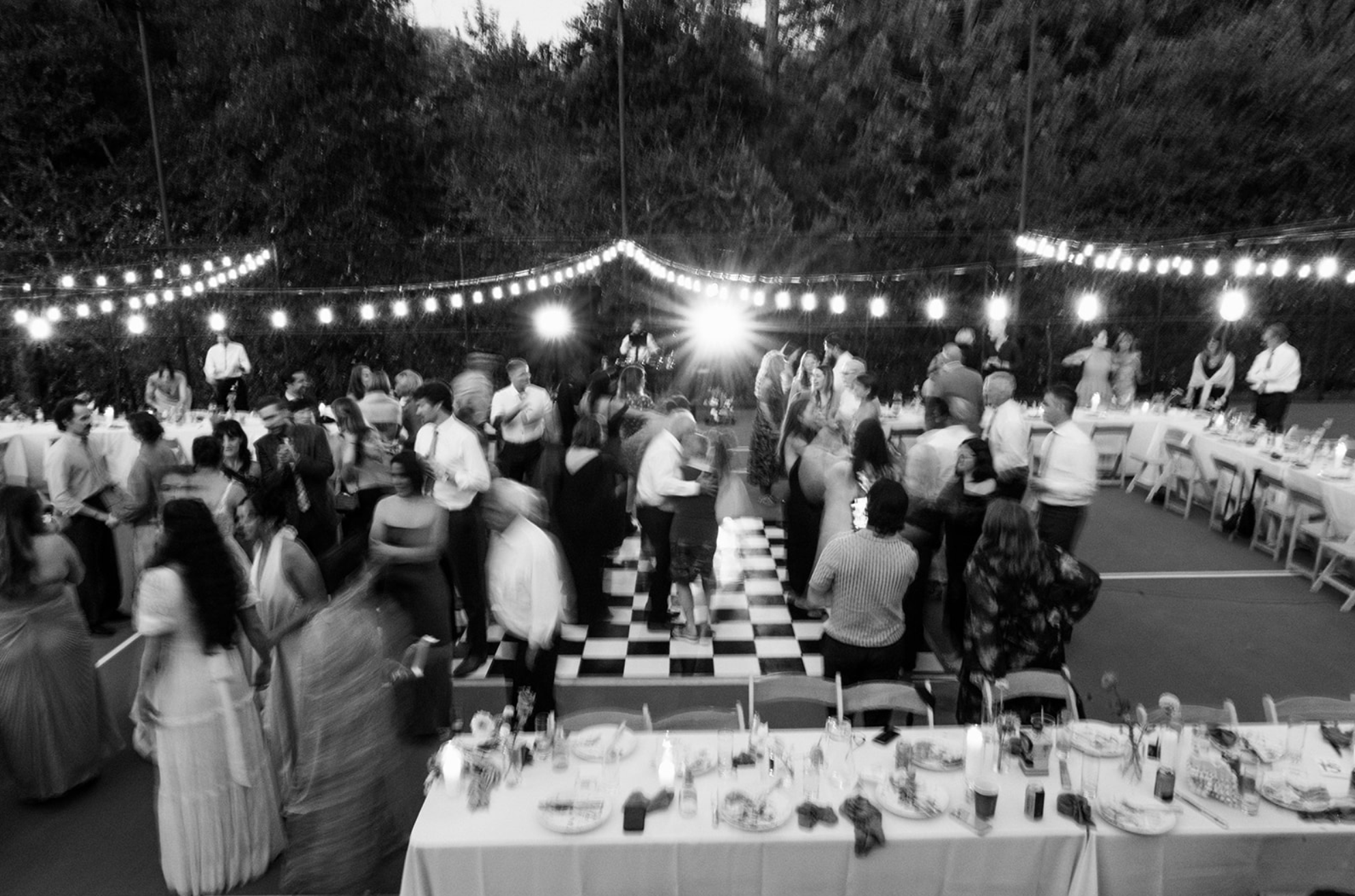People socializing and dancing at an outdoor evening event with string lights and a DJ, with a checkered dance floor and tables set for dining.