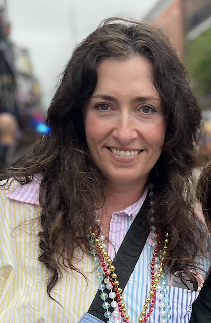 A woman with long, wavy brown hair smiling at the camera. She's wearing a light yellow and pink striped shirt with a collared pink shirt underneath. She has multiple Mardi Gras beads around her neck and a black strap across her chest. The background appears to be an outdoor street scene with blurred buildings and people.
