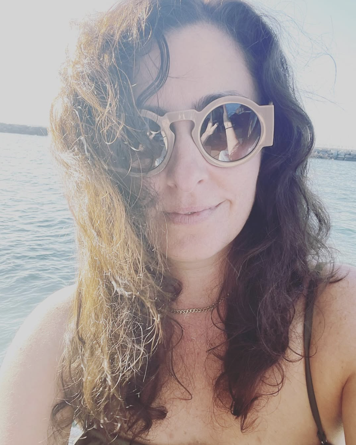 A woman with curly hair wearing sunglasses, smiling at the beach with water and a jetty in the background.