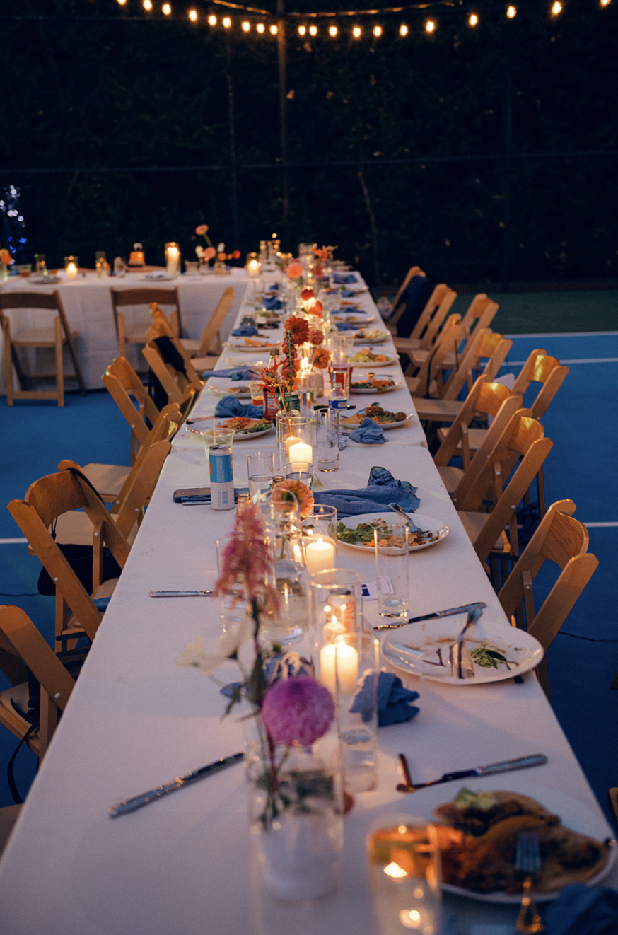 Long outdoor dinner table decorated with candles, flowers, and dinnerware, set for an evening celebration with string lights overhead.