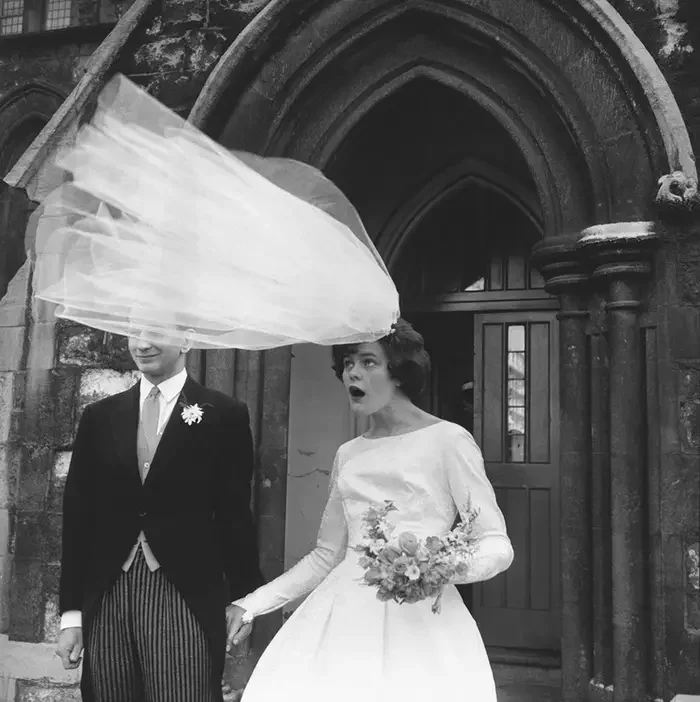Black and white photo of a bride and groom outside a gothic church. The bride, wearing a white dress and holding a bouquet, appears surprised or shocked. The groom, dressed in a formal suit with a boutonniere, stands beside her. The bride's veil is flying in the wind.