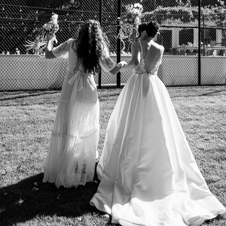 Two women in wedding dresses holding hands near a football field.