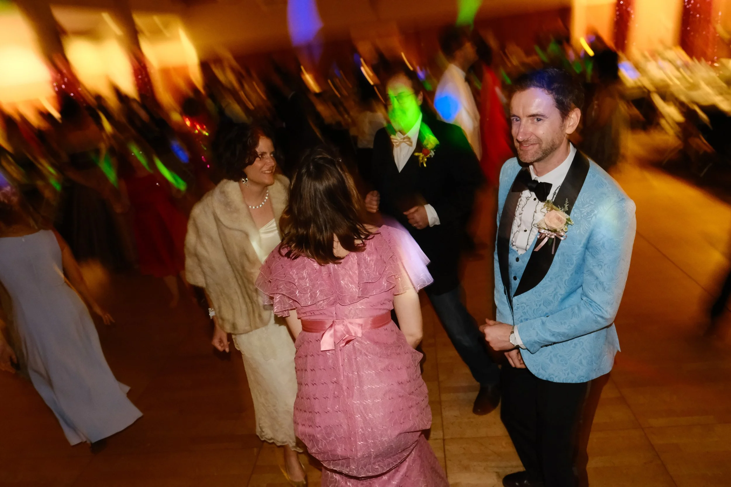 People dancing at a wedding reception, including a man in a light blue tuxedo with black lapels and a woman in a pink dress with ruffles.