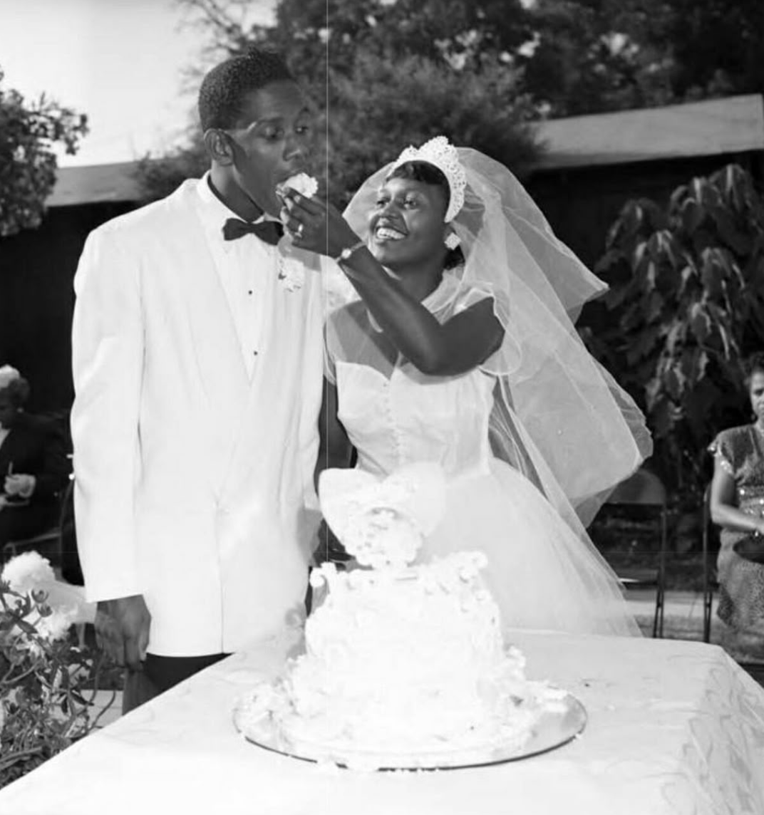 A bride playfully feeds cake to a groom at their wedding reception outdoors, with a wedding cake on the table and guests visible in the background.