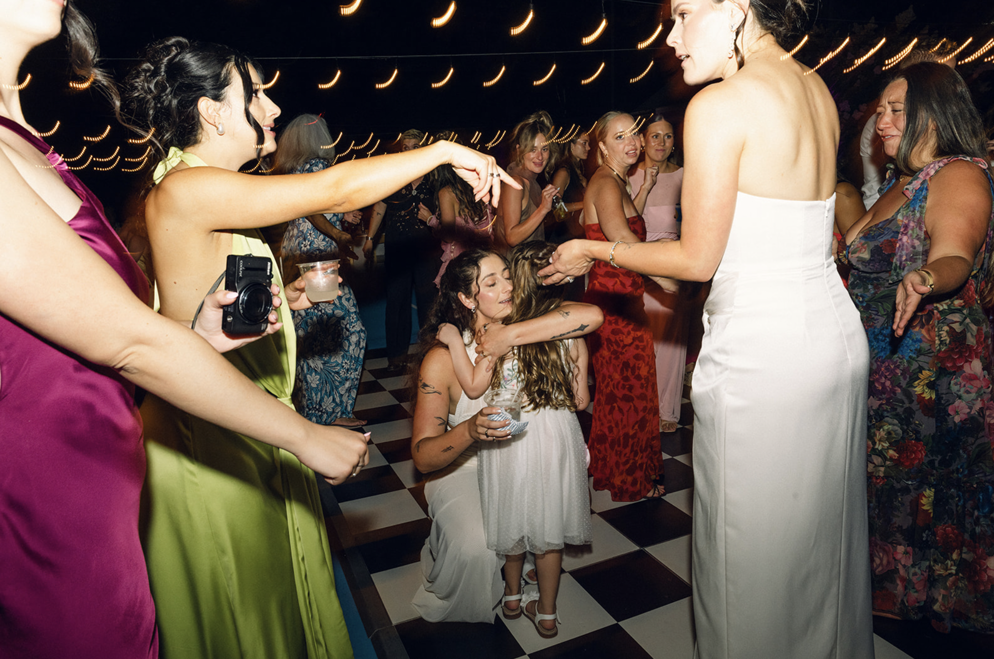 Group of women and children dancing and socializing at a lively indoor event with string lights overhead.