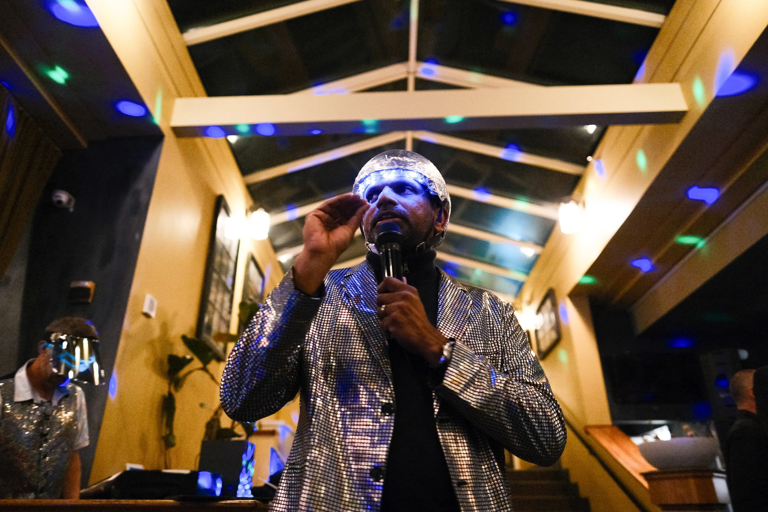 A man wearing a shiny, checkered blazer and a silver helmet performs with a microphone in hand in a dimly lit indoor space decorated with string lights and multicolored disco lights.
