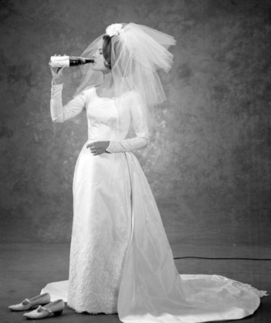 Black-and-white photo of a bride in a wedding dress, wearing a veil, drinking from a bottle.