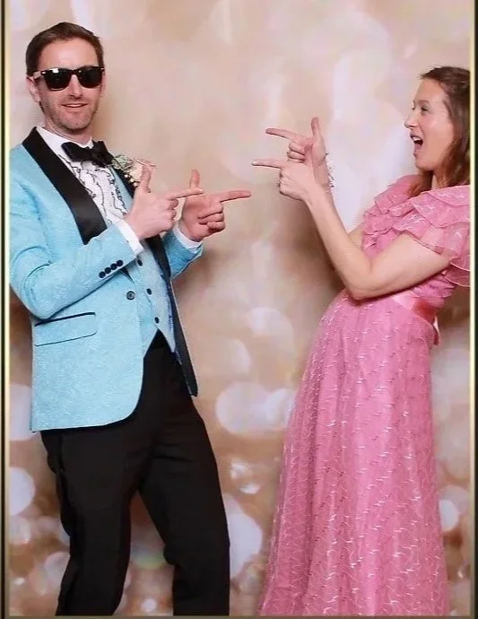 A man in sunglasses wearing a light blue tuxedo jacket and a woman in a pink dress pointing at each other and laughing in front of a neutral, bokeh background.