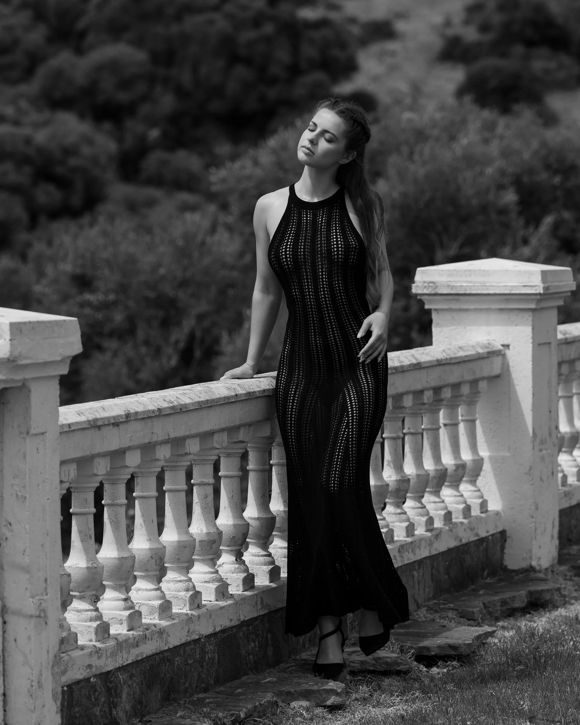 A woman in a long, sleeveless, patterned dress standing outdoors by a stone balustrade fence with mountains and trees in the background.