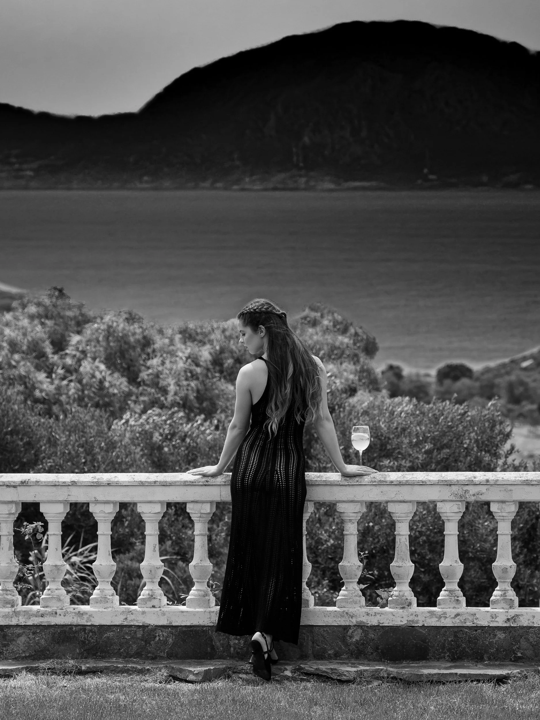 A woman in a black dress and high heels standing on a balcony with a glass of wine. She is looking down with her hands resting on the railing. The background features a scenic view of a large hill or mountain across a body of water, with lush greener