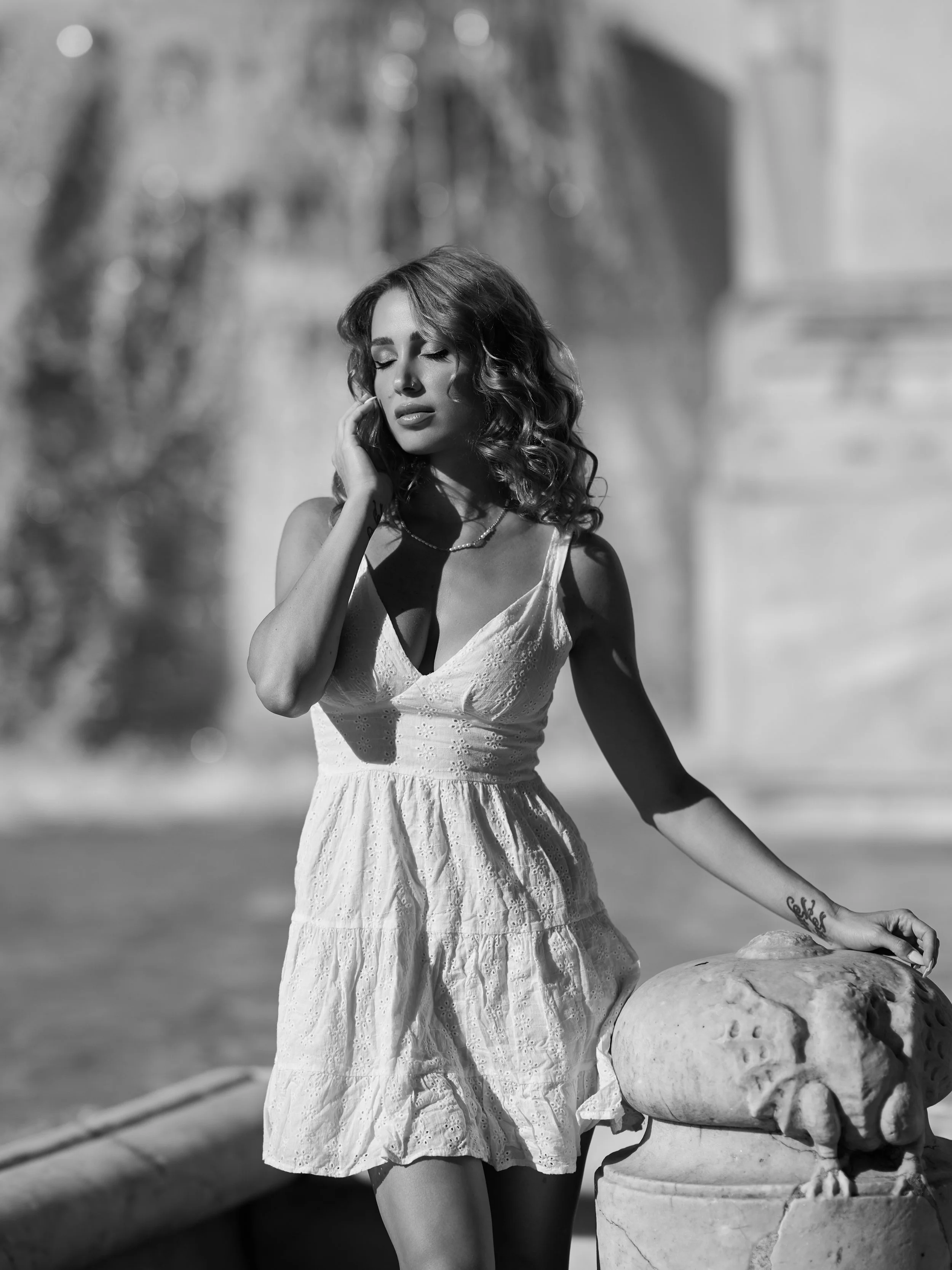 A young woman with curly hair, wearing a light-colored dress, stands outdoors next to a stone lion sculpture on a pedestal, with her hand touching her face and eyes closed.