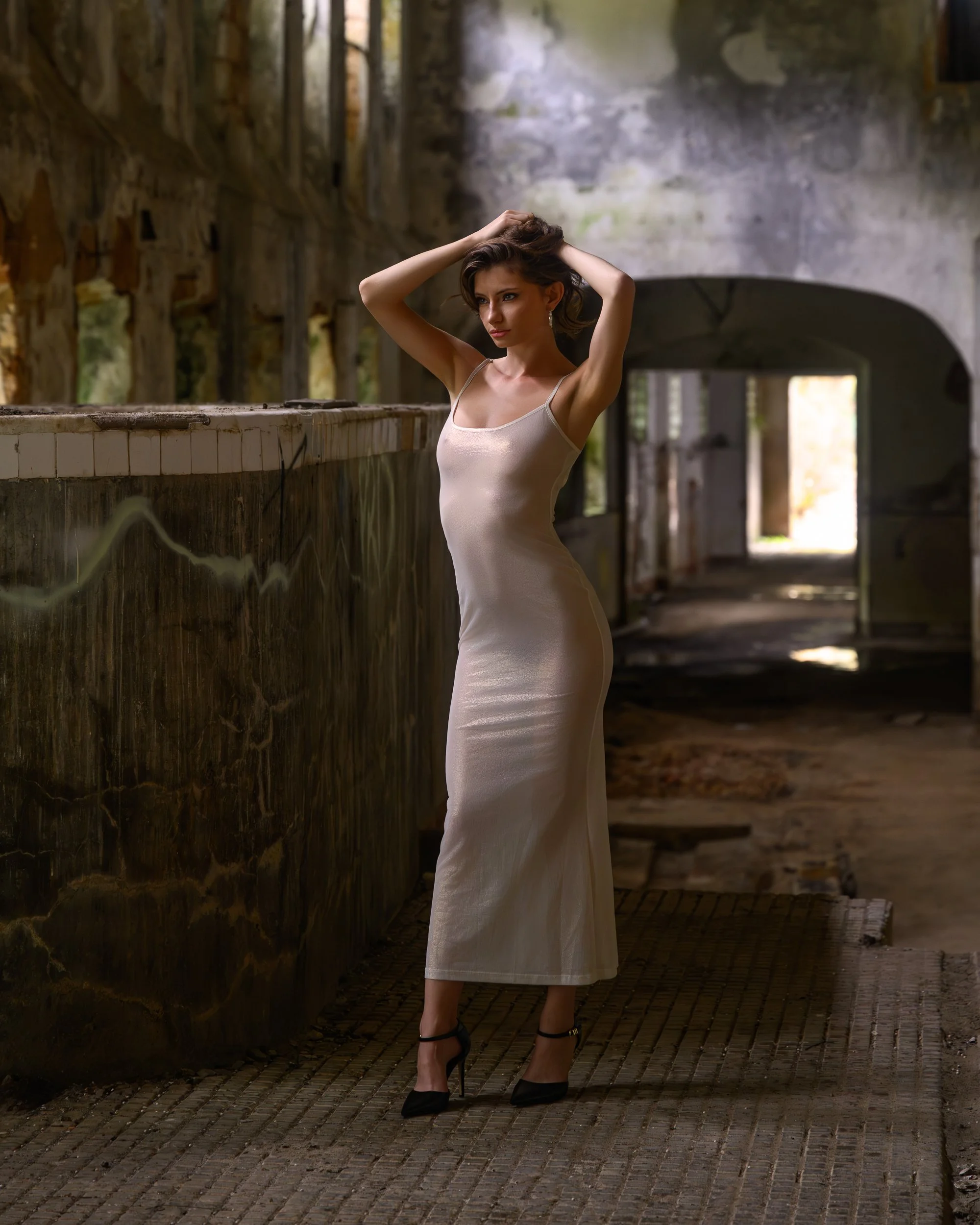 A woman in a beige dress and black high heels standing in an old, abandoned building with stone walls and a brick floor, posing with her hands in her hair.