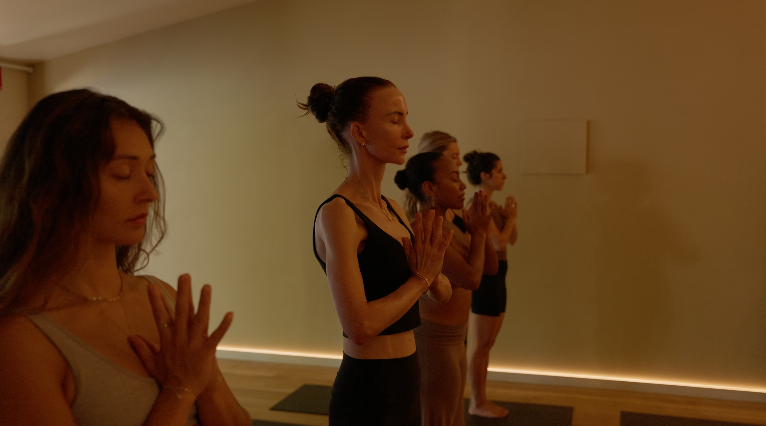 Group of women practicing meditation or yoga in a calm, dimly lit room, standing with eyes closed, hands in a prayer position.
