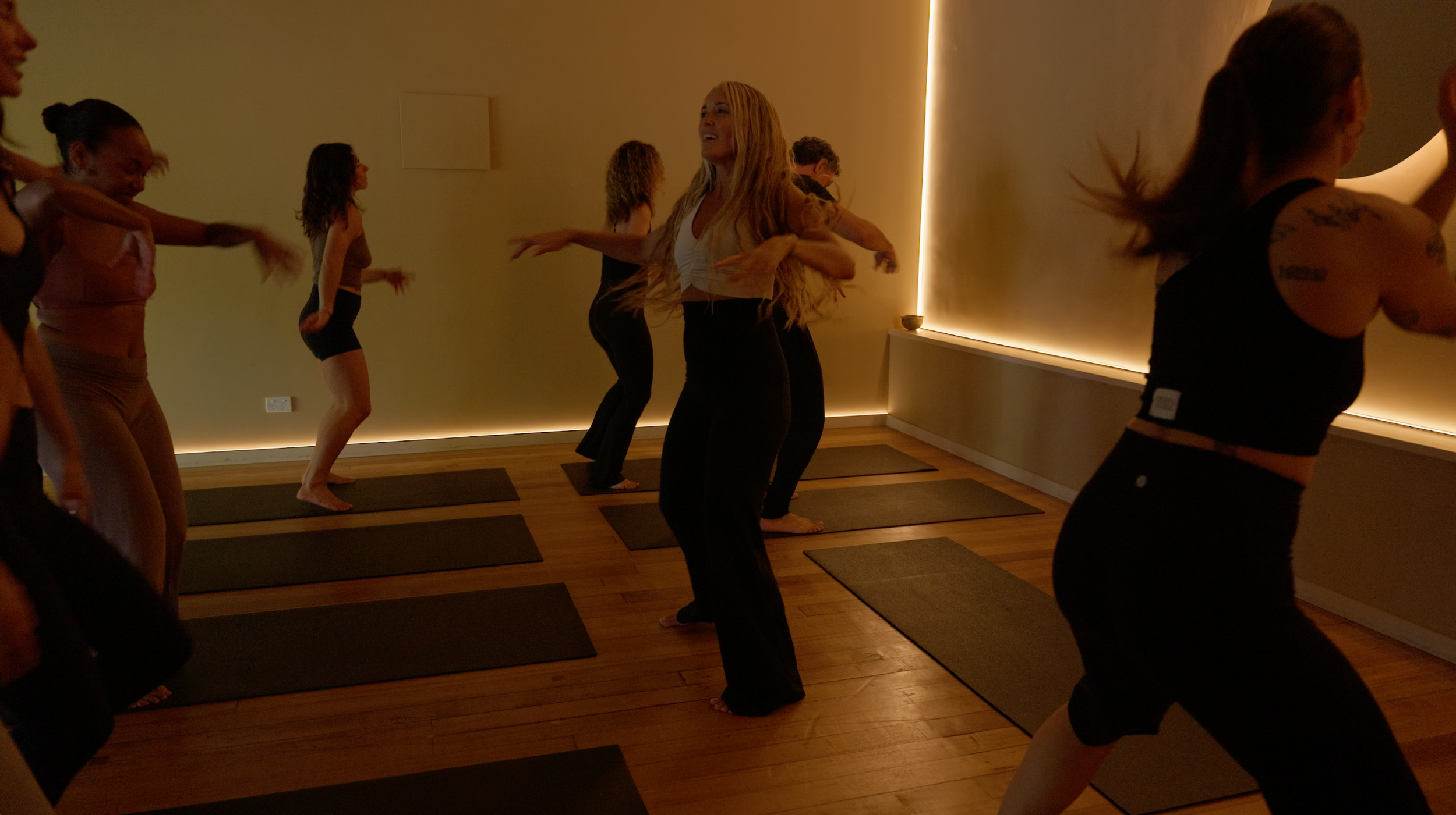 Group of women doing yoga in a dimly lit room with wooden floors and mats, some smiling and stretching.