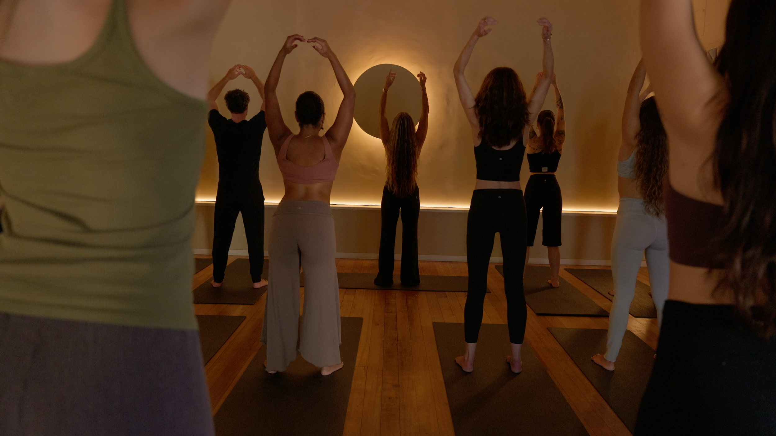 A group of people participating in a yoga or meditation class in a dimly lit room, standing on yoga mats, with some raising their hands overhead.