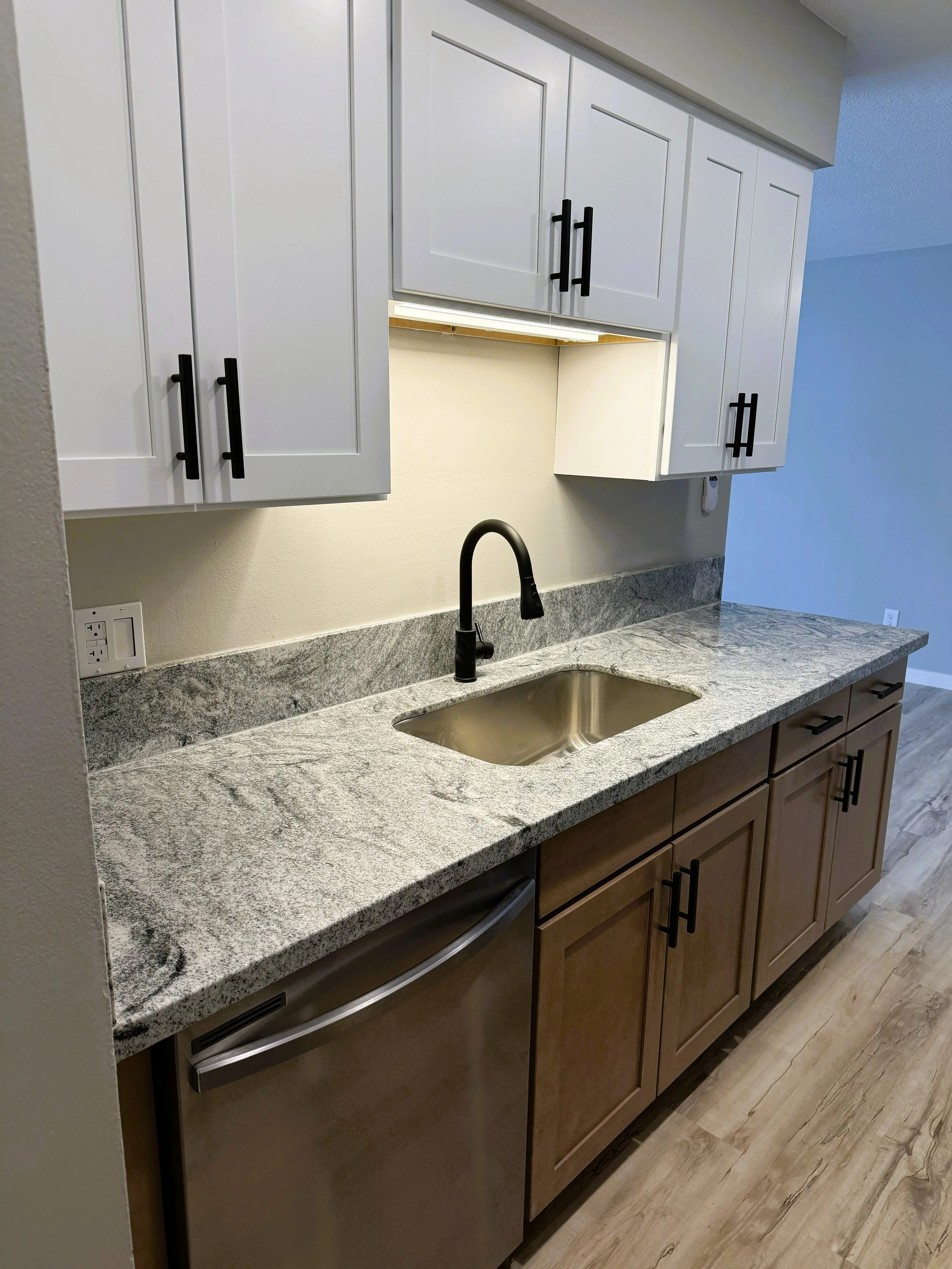 A kitchen countertop with a granite surface, a stainless steel sink, a black faucet, and wooden cabinets with black handles.