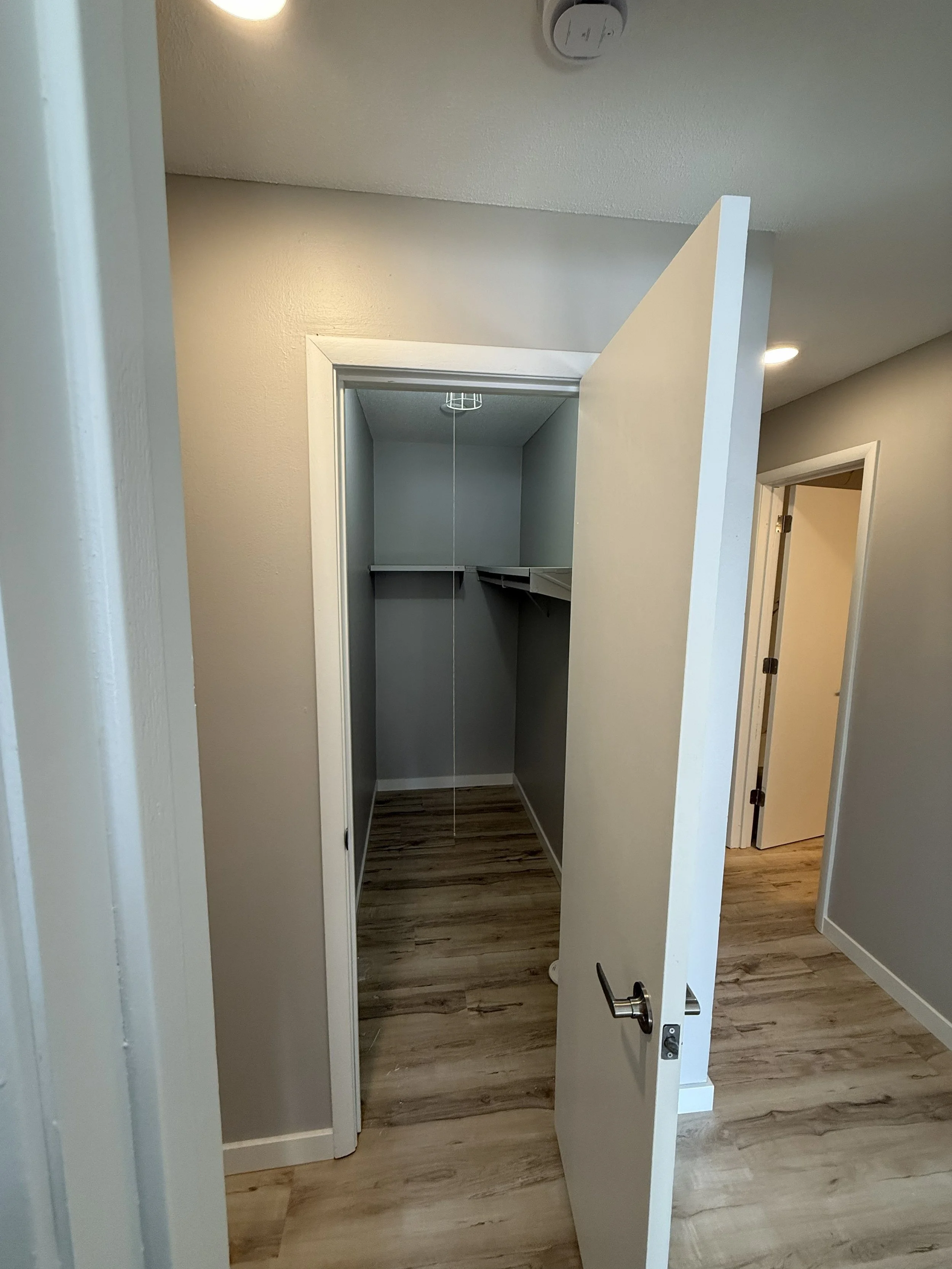 An open closet with a bare interior, white trim, and a mirror on the inside of the door, standing on wood flooring in a room with beige walls and ceiling lights.