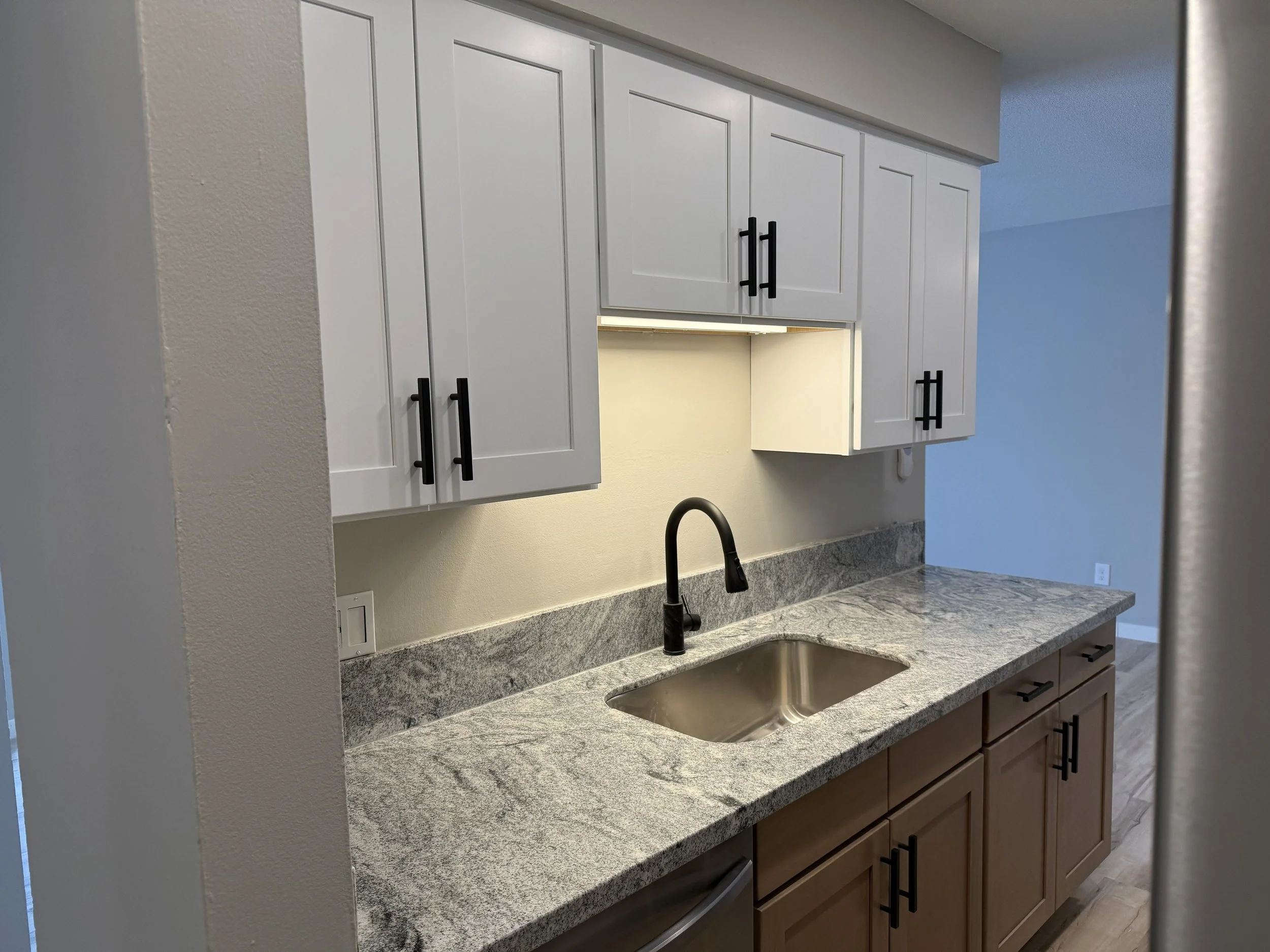 Kitchen with white upper cabinets, brown lower cabinets, granite countertop, and a black faucet over a stainless steel sink.