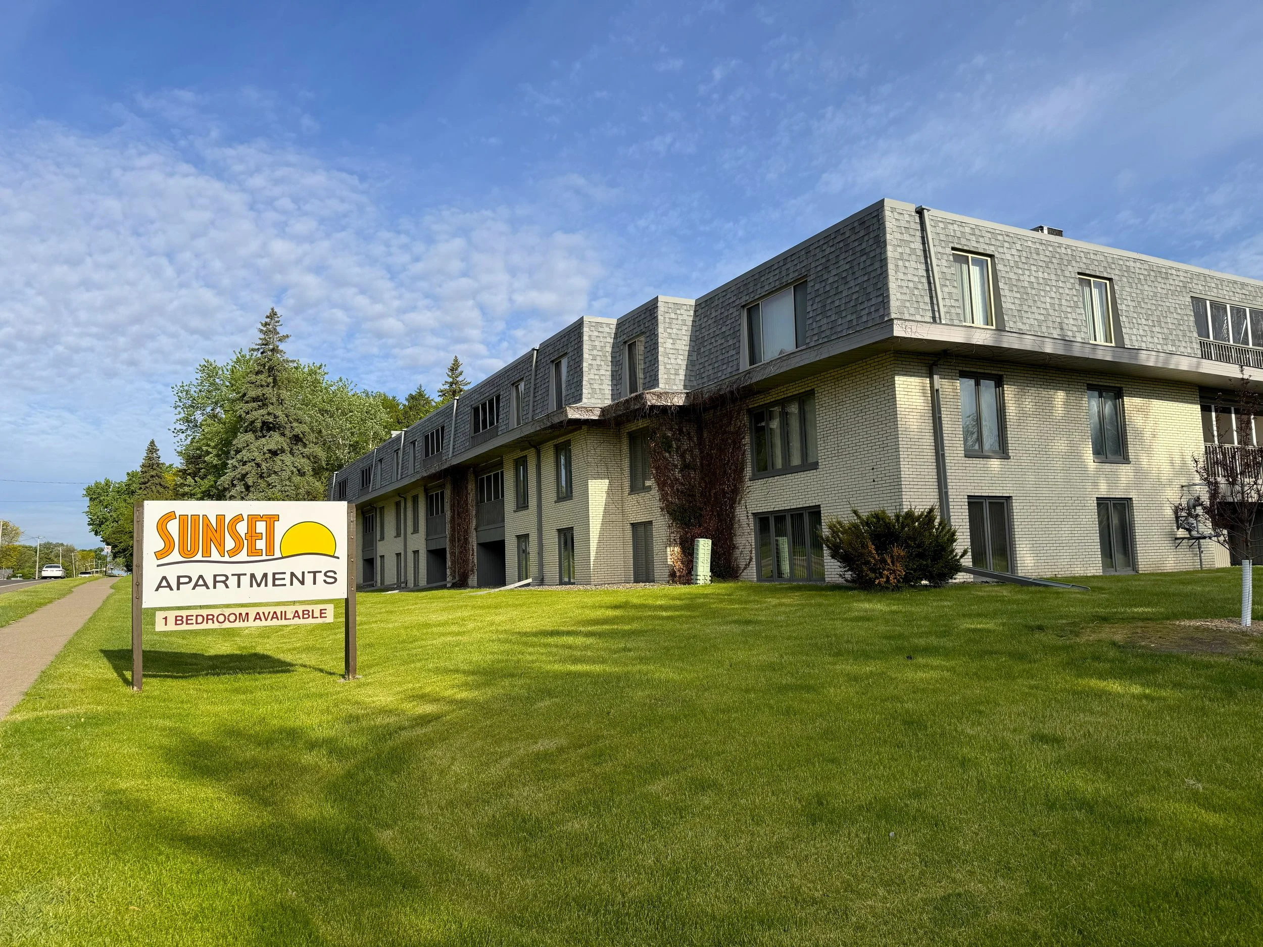 A multi-story residential building with a gray shingle roof, cream brick walls, and several large windows. There is a lawn with trees and bushes in front of the building. A sign in the foreground reads "Sunset Apartments" and offers a one-bedroom ava