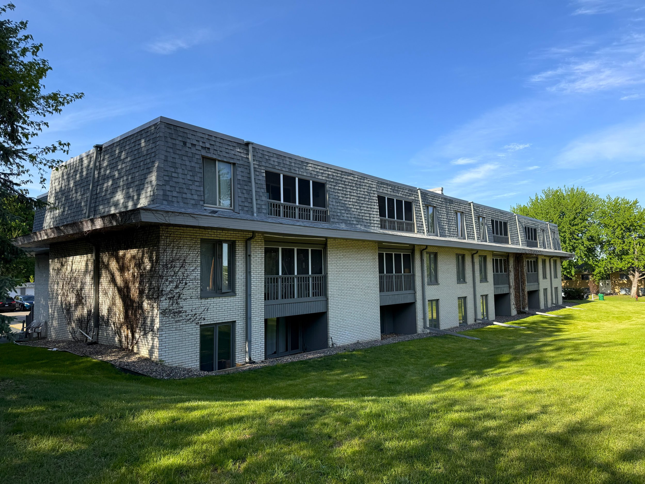 A multi-story residential building with a brick lower level and a wooden-shingled upper level, set against a bright blue sky with some clouds and green trees nearby.