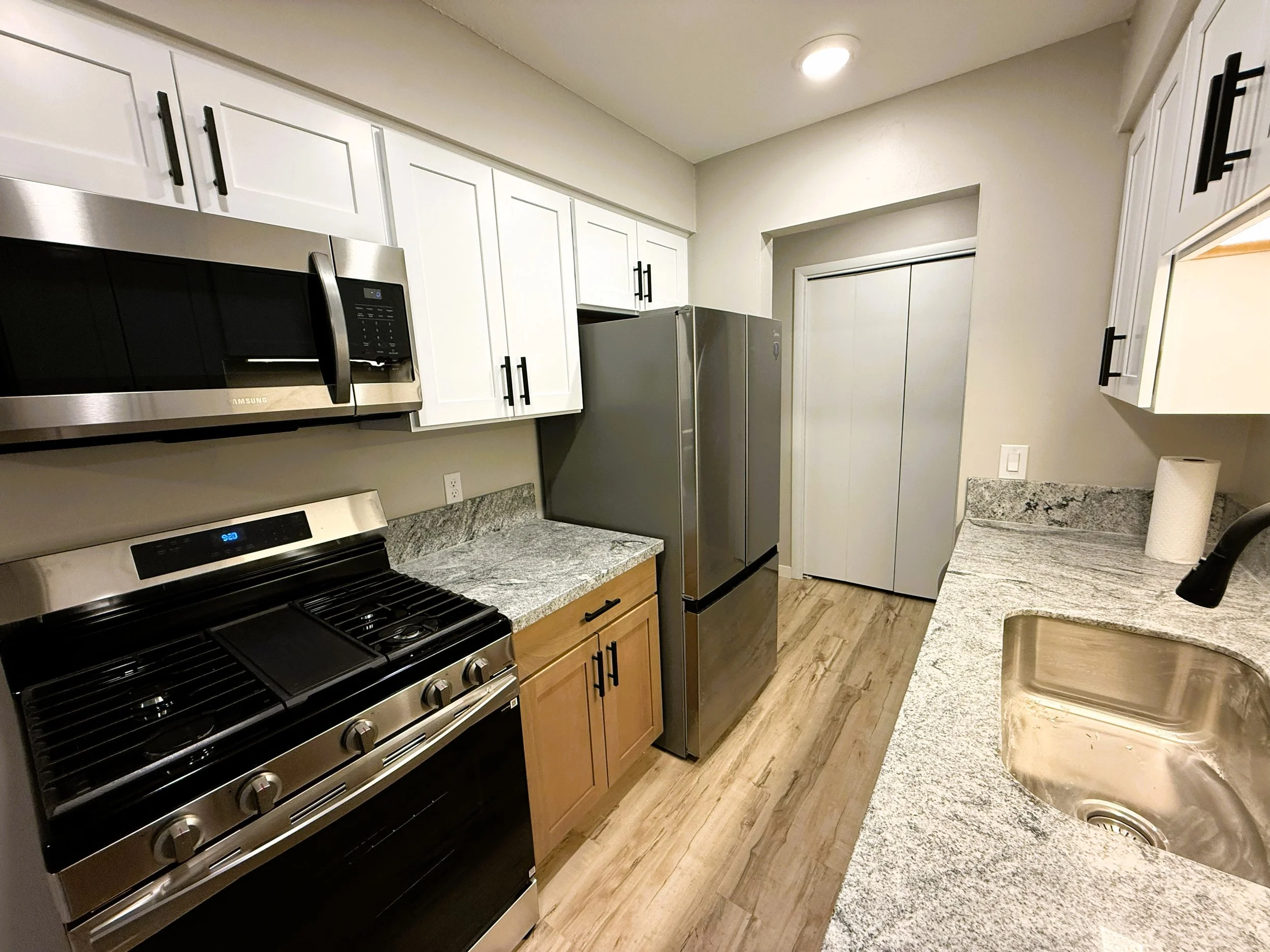 A modern kitchen with white upper cabinets, black handles, a stainless steel microwave, a stainless steel refrigerator, a gas stove, granite countertops, and wood flooring.