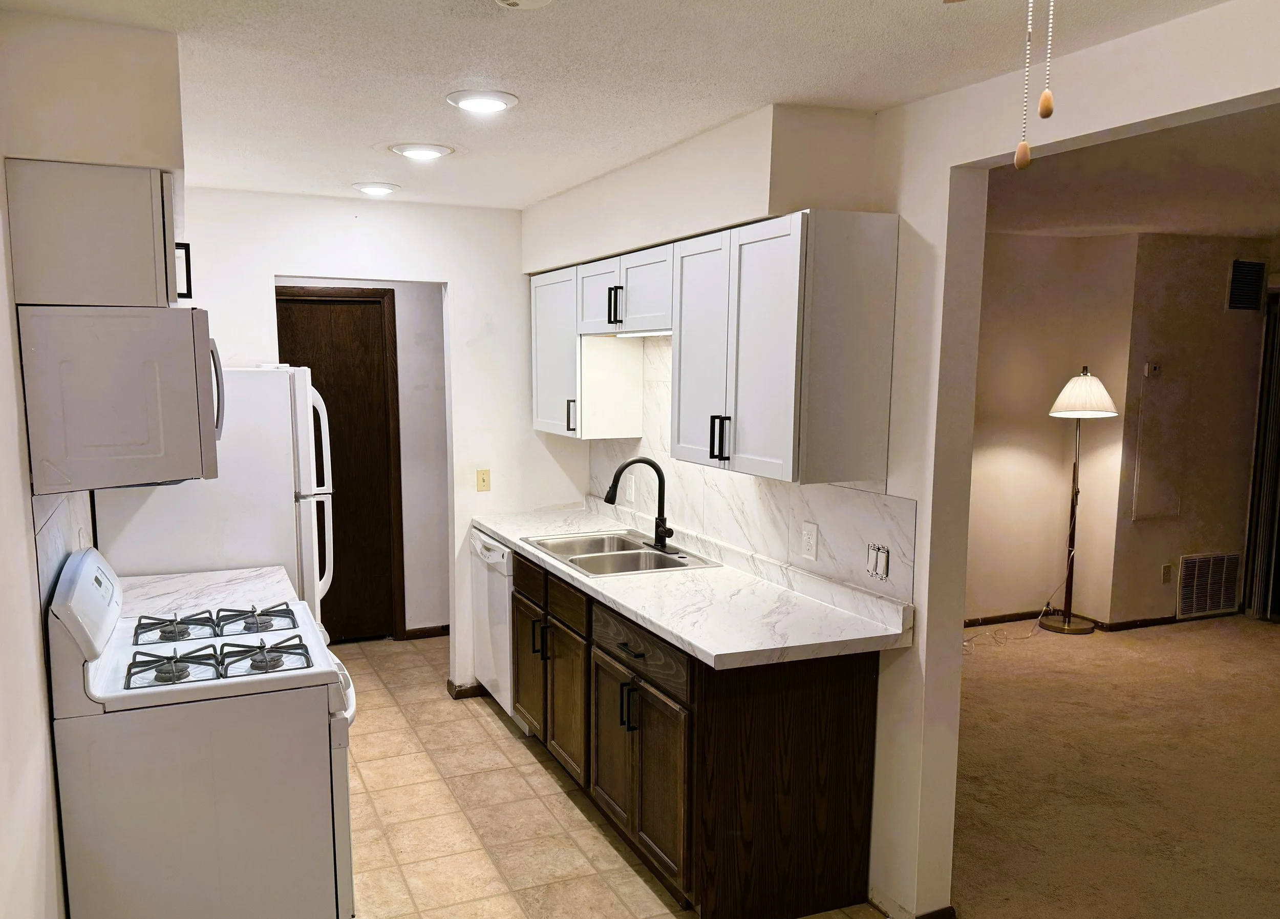 A kitchen with white upper cabinets, dark lower cabinets, a marble countertop, a double sink, a white stove, and white refrigerator. There is a living area to the right with a floor lamp. Recessed lighting is installed in the ceiling.