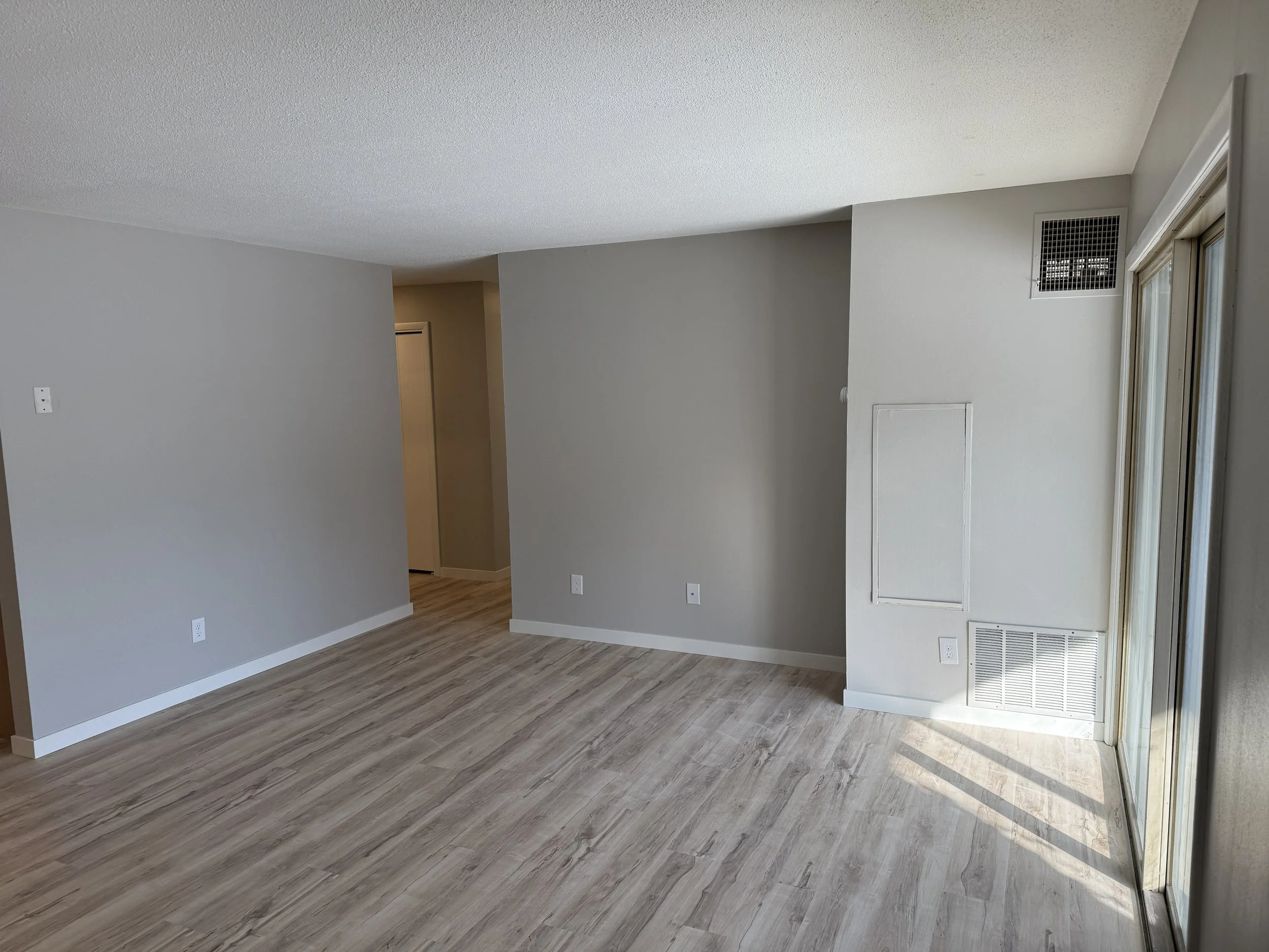 Interior view of a modern room with light-colored walls, wood flooring, and a sliding glass door to the right. There is an air vent above the door and several electrical outlets along the walls.