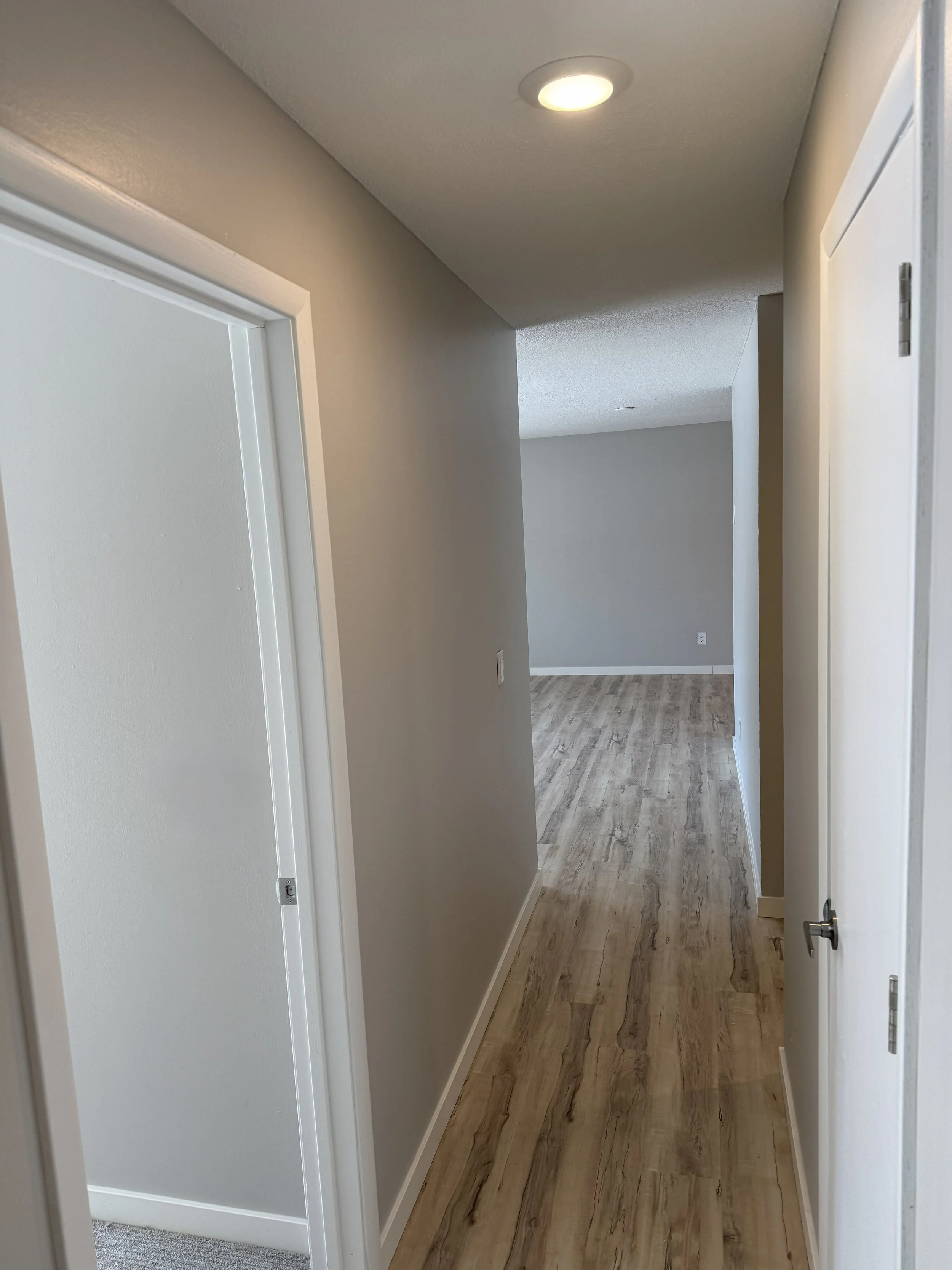 A hallway with beige walls, wooden flooring, ceiling light, several doorways, and a view into another room.
