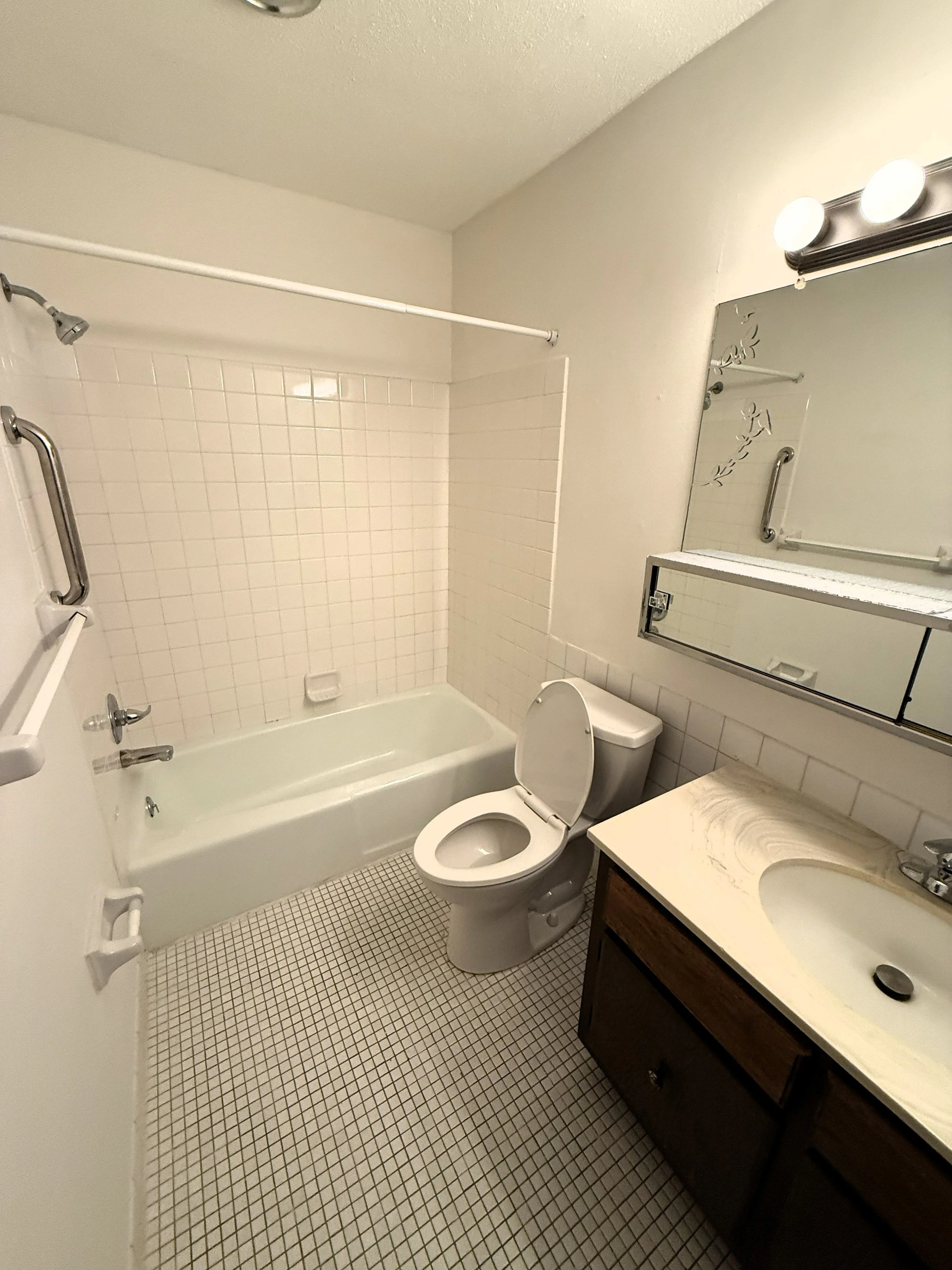A small bathroom with white tile flooring and a shower/tub combination with a grab bar. There is a white toilet next to a dark wood vanity with a beige marble countertop and oval sink. A mirror with mounted lights is above the sink, and a towel rod i