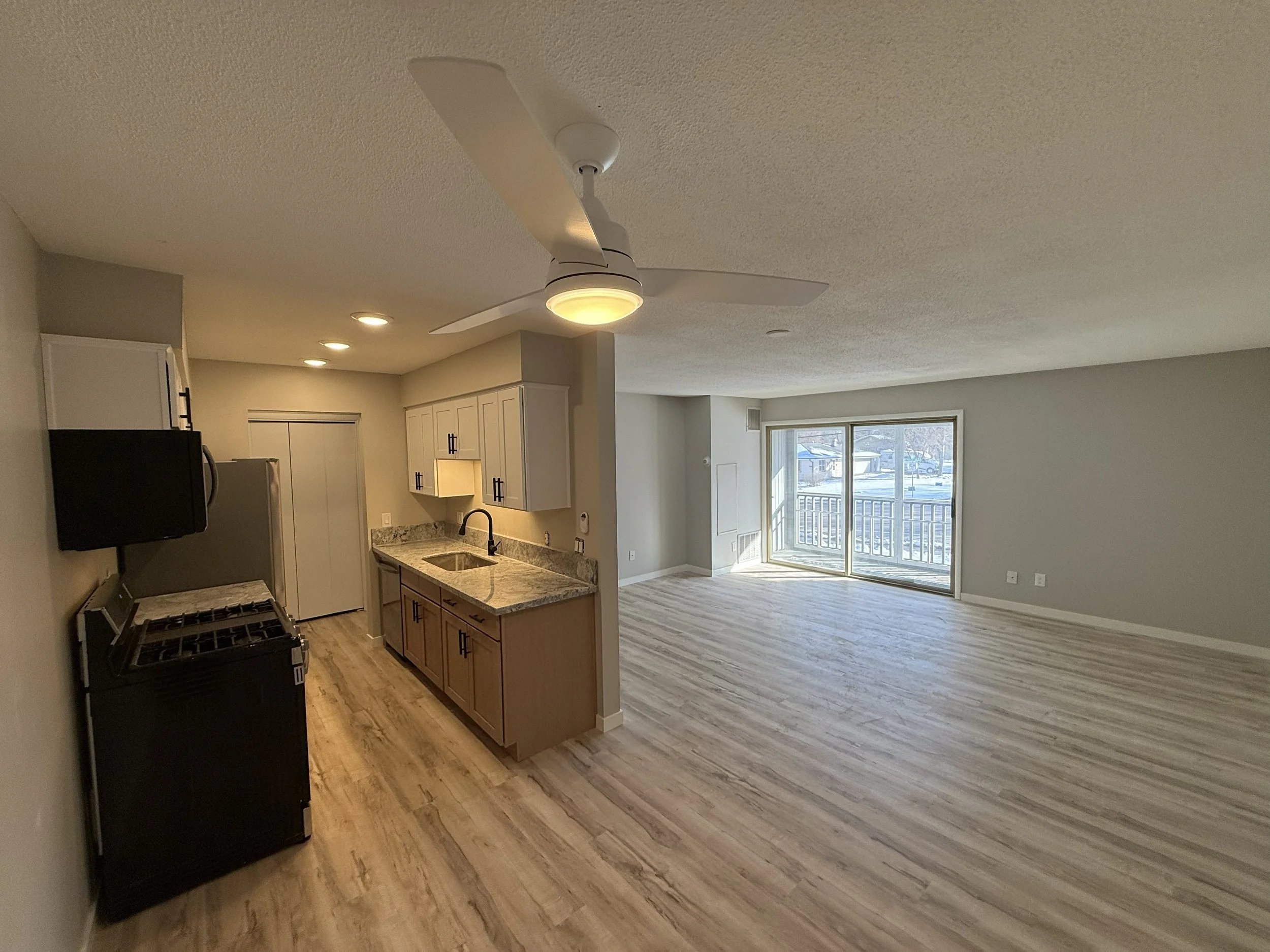 An open-concept kitchen and living room with wood flooring, white cabinets, a black refrigerator, and a sliding glass door leading to a balcony, letting in natural light.