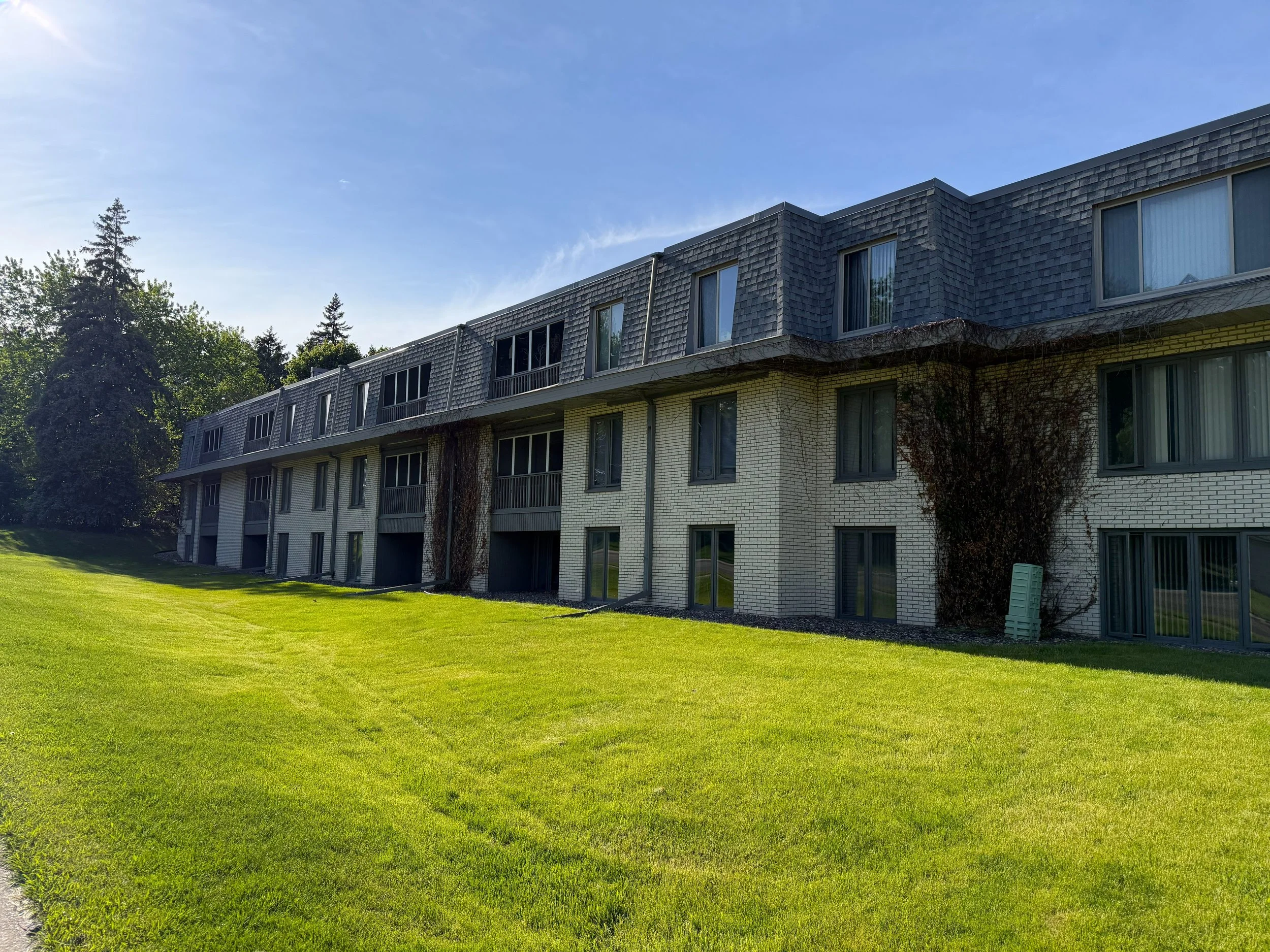 A multi-story apartment building with a brick and shingle exterior, large windows, and small balconies, surrounded by a green lawn and trees under a clear blue sky.