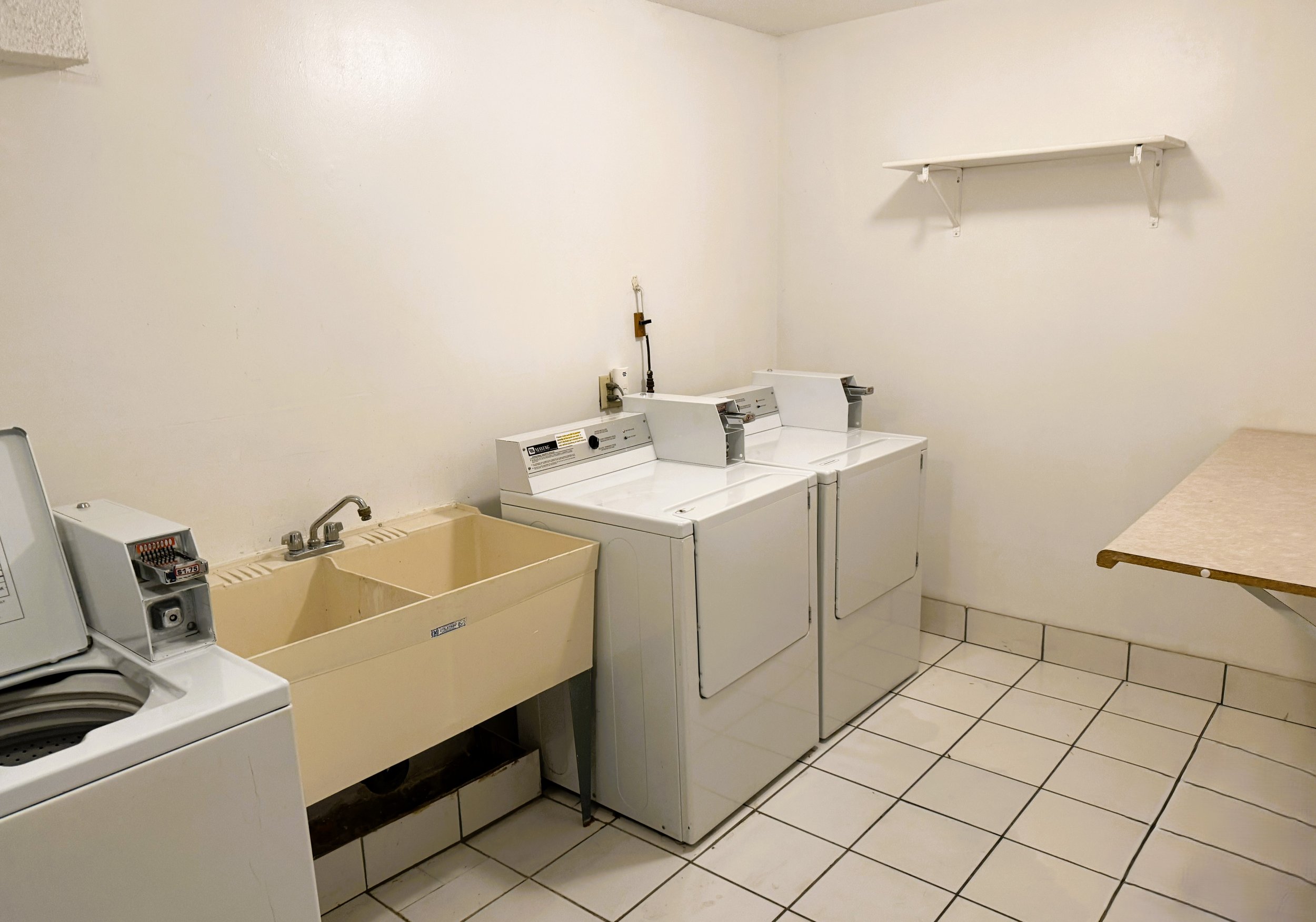 Laundry room with a beige utility sink, a beige washing machine, and two white dryers against a cream-colored wall and tiled floor, with a white shelf on the wall.