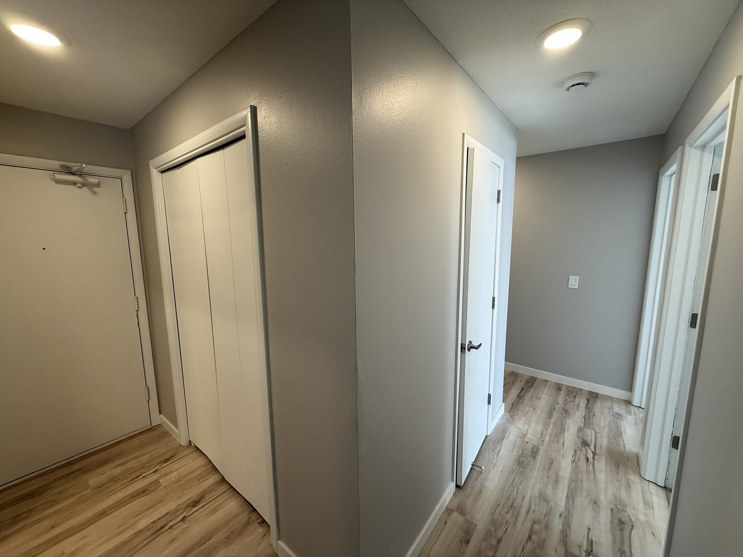 A hallway with white walls and doors, one of which is a closed door with a door closer, and another door slightly open, revealing a bit of the room beyond. The hallway has wood flooring and recessed ceiling lights.