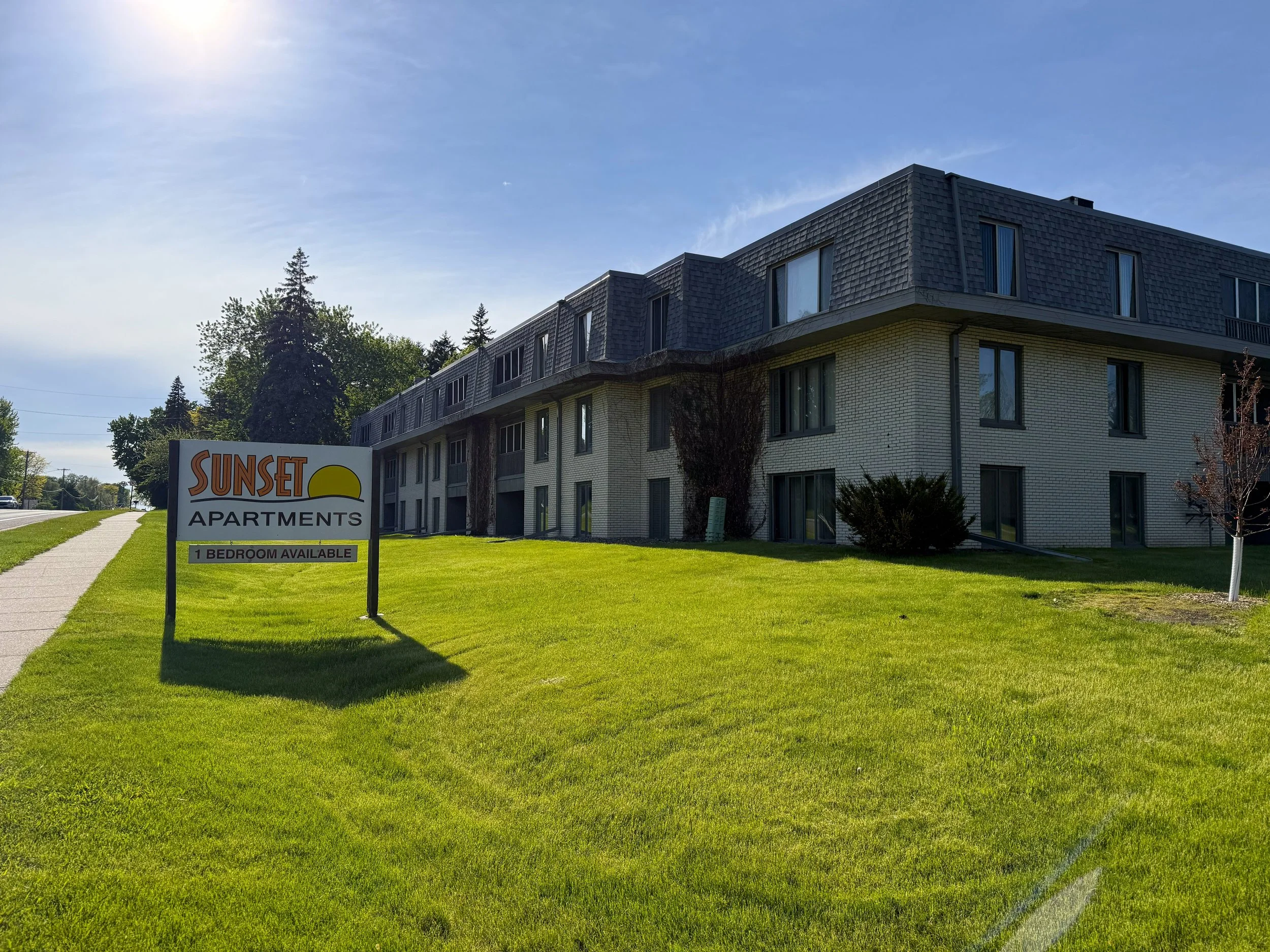 A multi-story apartment building with a white brick exterior and dark gray shingle roof, situated next to a sidewalk and green lawn with a sign that reads "Sunset Apartments" indicating one bedroom available. There are trees in the background against a clear blue sky with some wispy clouds.