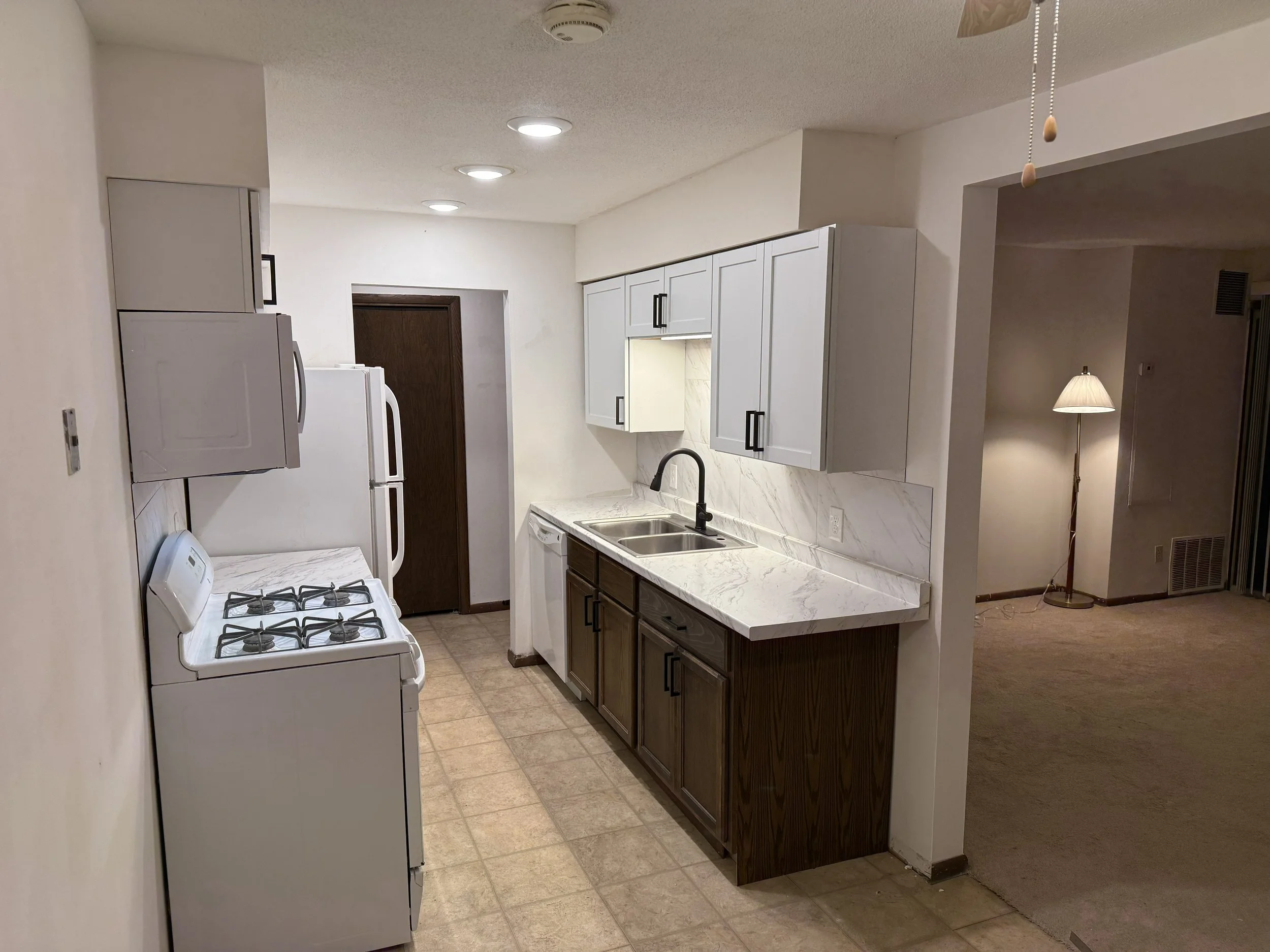 Kitchen with white cabinets and appliances, a marble countertop with a sink, and a beige floor. There is a doorway leading to another room with a standing lamp.