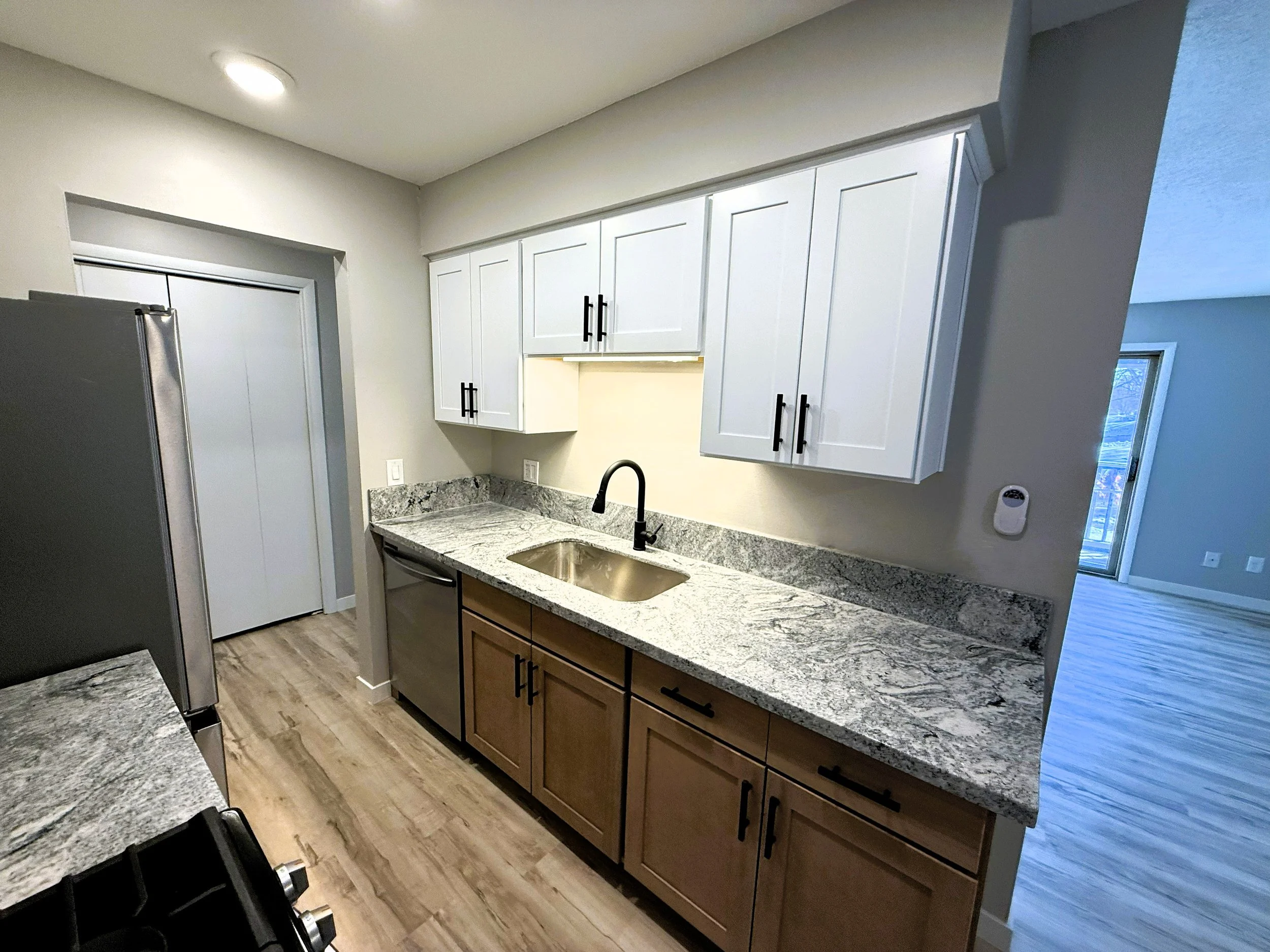 Kitchen with granite countertops, white upper cabinets with black handles, brown lower cabinets, stainless steel refrigerator, sink with black faucet, and wooden flooring.