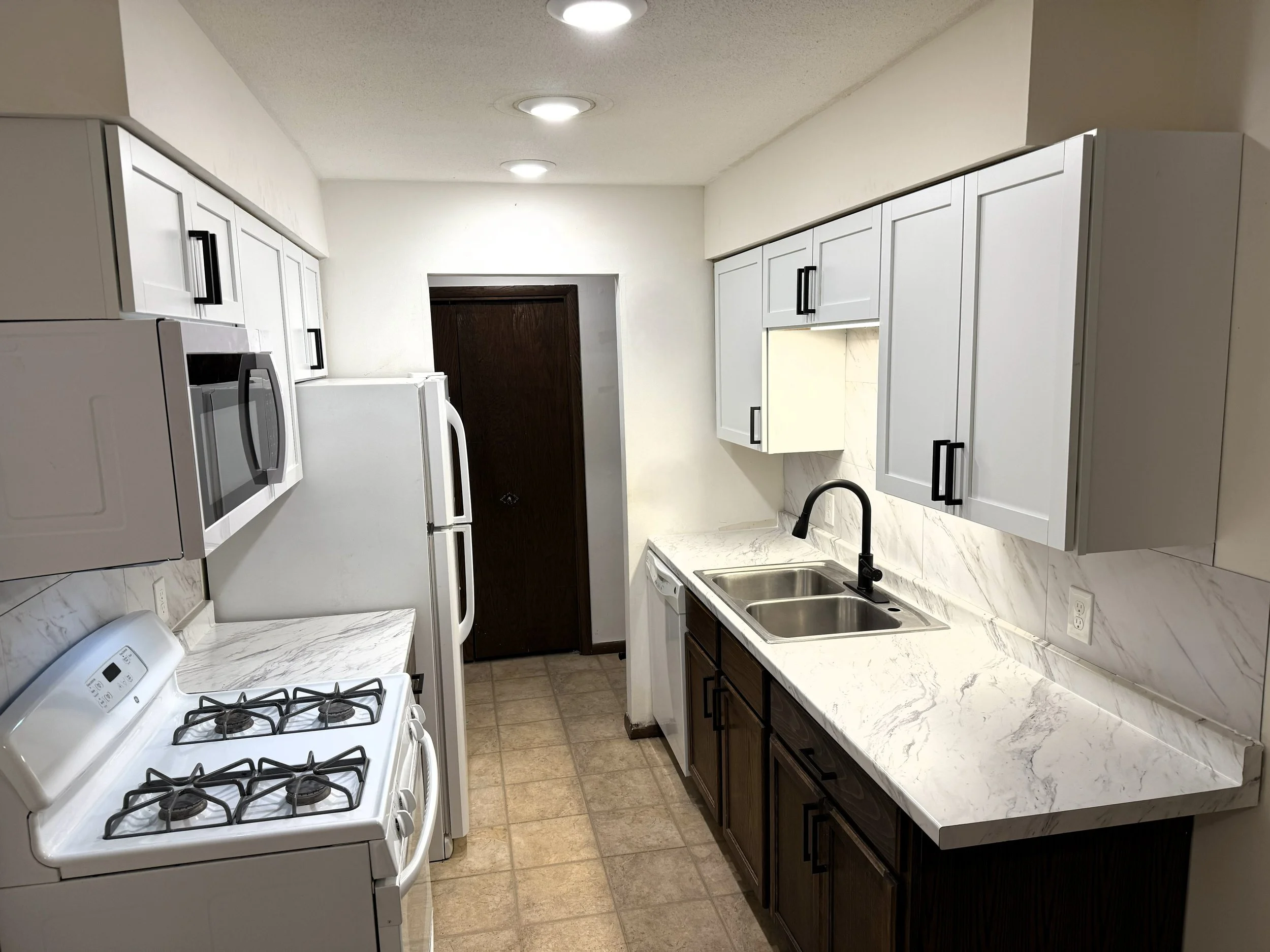 A small kitchen with white countertops, white cabinets with black handles, a white gas stove, and a double-basin stainless steel sink. There is a white refrigerator next to the stove and a microwave on the counter. The walls are off-white, and the fl
