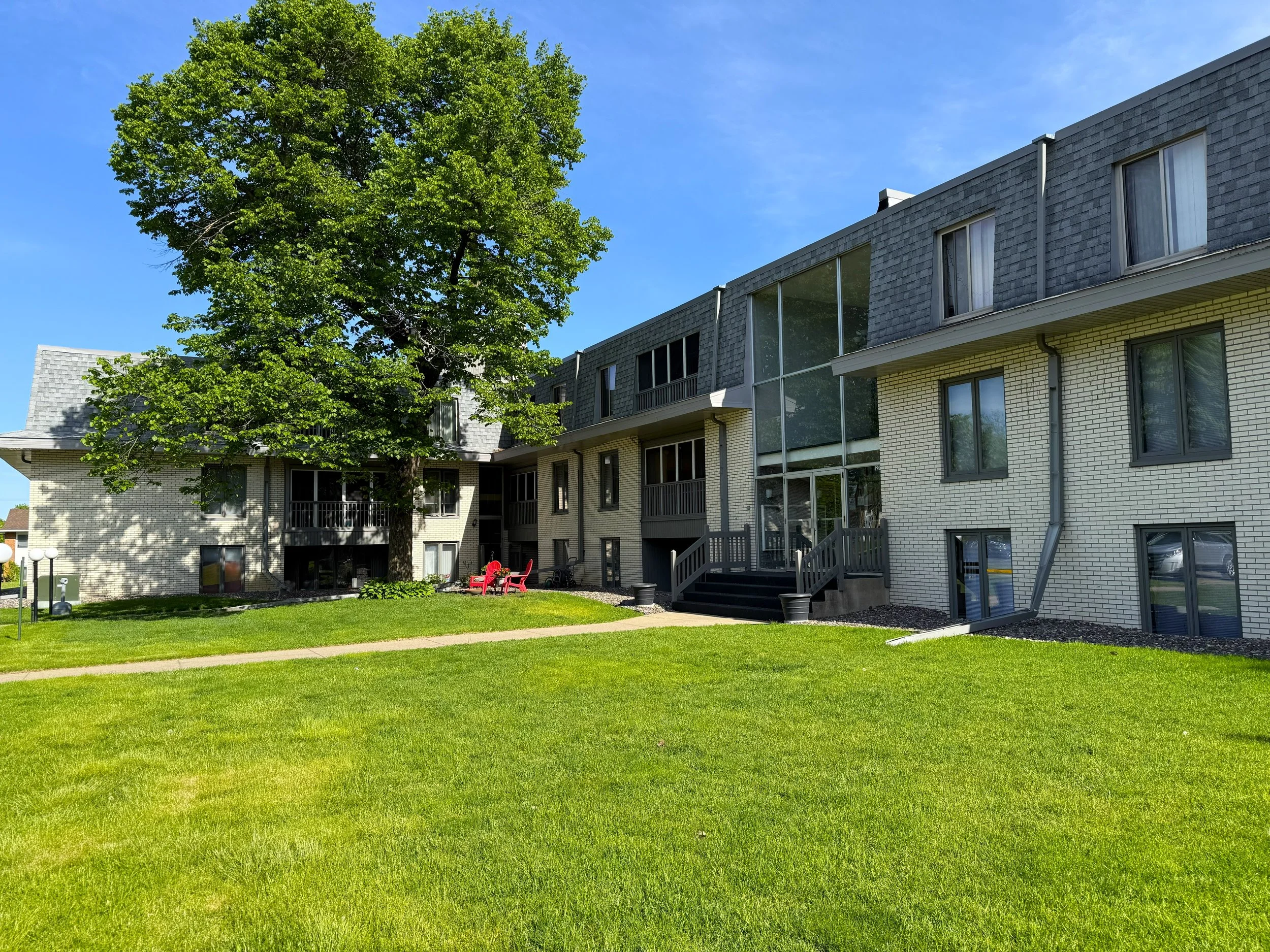 A residential apartment complex with multiple levels, featuring yellow brick walls, large windows, and balconies. A large green tree is in the foreground, with a clear blue sky overhead and a well-maintained grassy lawn.