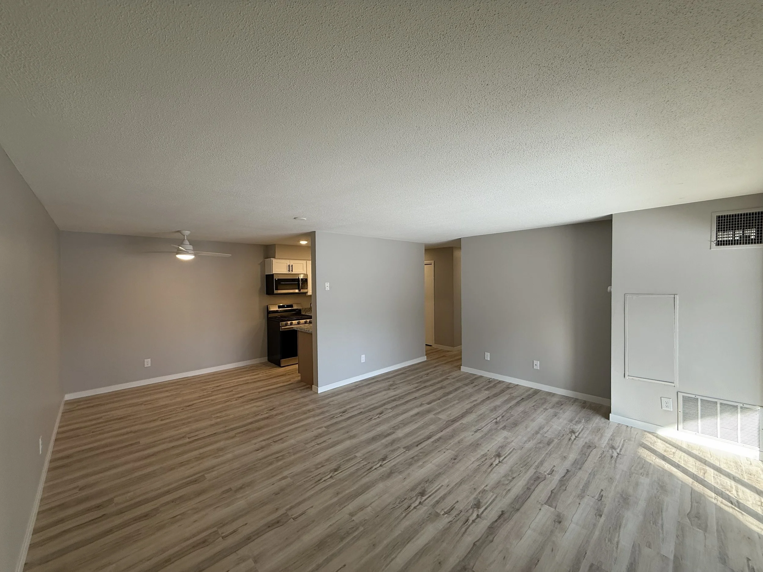 An open floor plan living area with wood laminate flooring, light gray walls, a kitchen in the background with white cabinets and black appliances, a ceiling fan, and a vent on the wall.