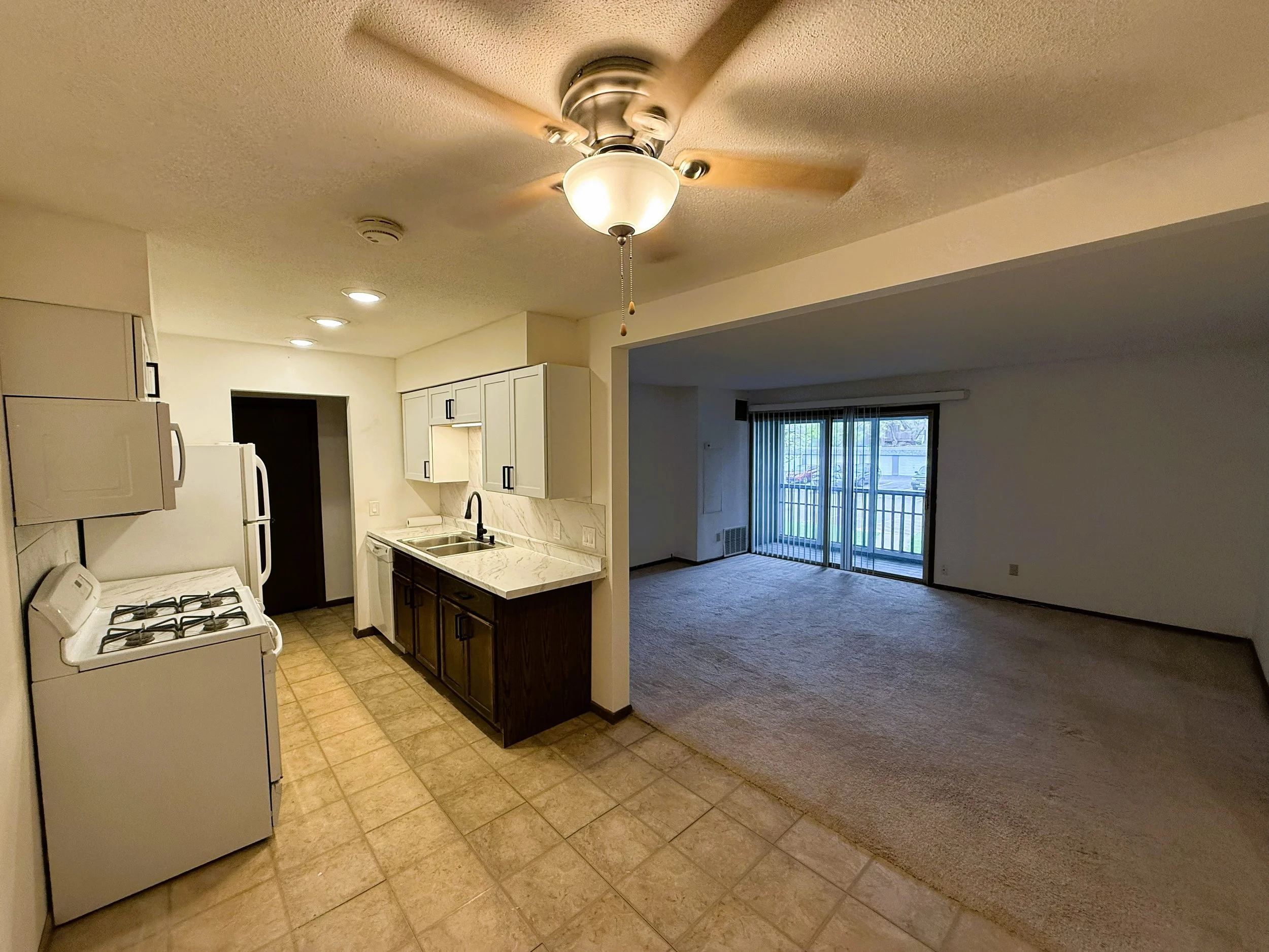 View of a kitchen with white cabinets, a white stove, and a refrigerator, adjacent to a living area with sliding glass doors and vertical blinds.
