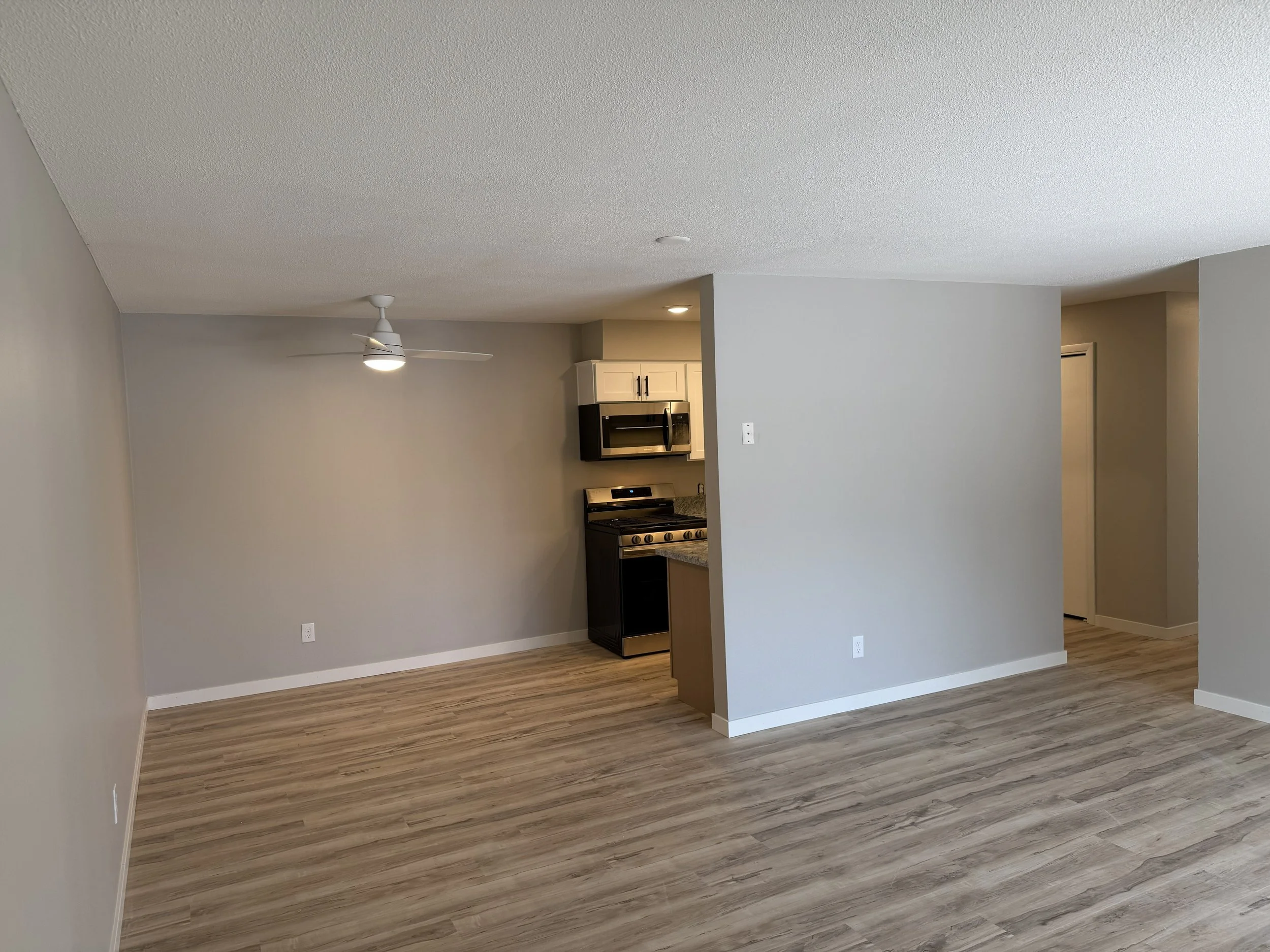 Interior view of an open living space with a kitchen area, beige walls, a ceiling fan, wood-style flooring, and a partial wall dividing the living and kitchen areas.